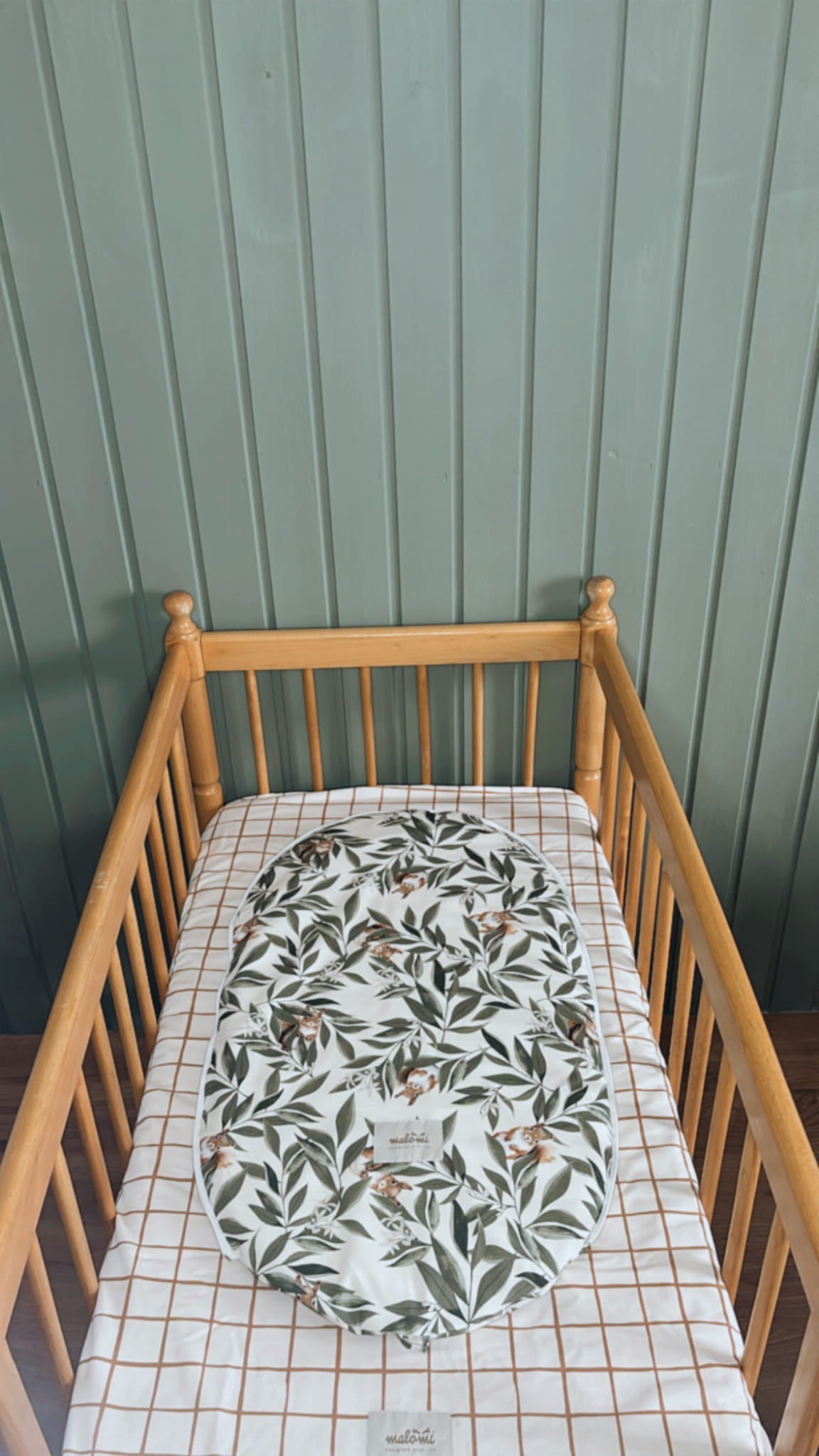 Babymatrasbeschermer met woodlandprint in een houten ledikantje met geruit hoeslaken. De beschermer is gemaakt van katoen en toont een speels motief van blaadjes en eekhoorns.