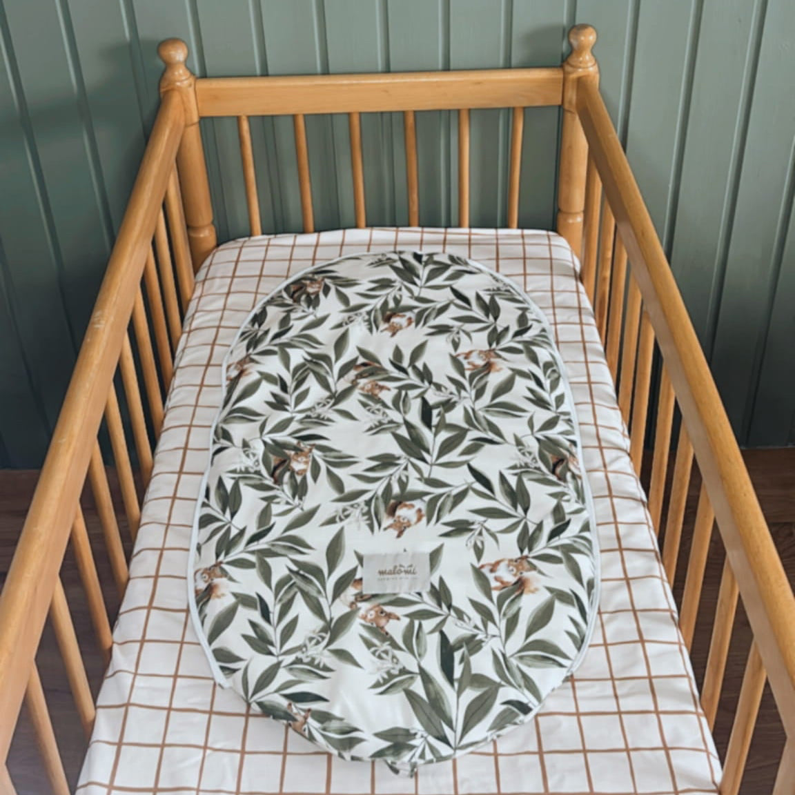 Babymatrasbeschermer met woodlandprint in een houten ledikantje met geruit hoeslaken. De beschermer is gemaakt van katoen en toont een speels motief van blaadjes en eekhoorns.