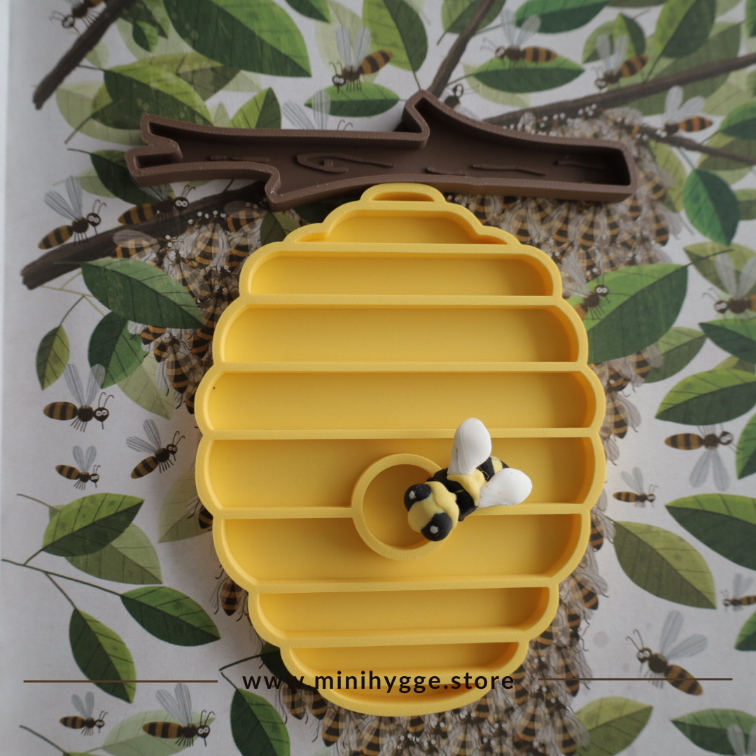 Gele bijenkorfvormige sensorische speel tray met compartimenten, vergezeld door een kleine handgemaakte bij en een bruine boomtak accessoire, geplaatst tegen een natuur-geïnspireerde achtergrond met geïllustreerde bijen en bladeren. De website "www.minihygge.store" wordt onderaan weergegeven.