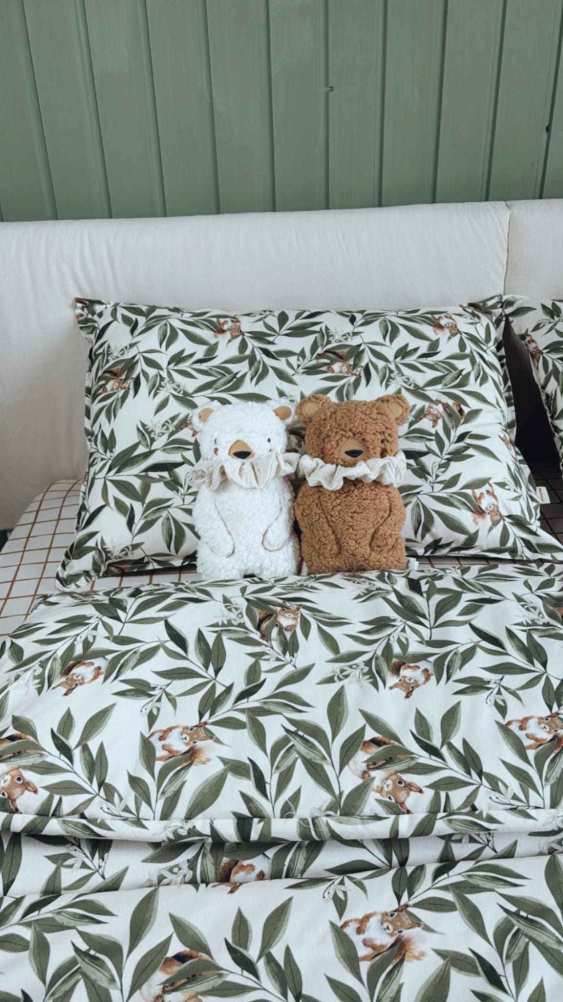 Twee pluche beren met ruches om hun nek zitten op een bed met beddengoed bedrukt met bladeren en eekhoorns. De achtergrond is een groen houten paneel.