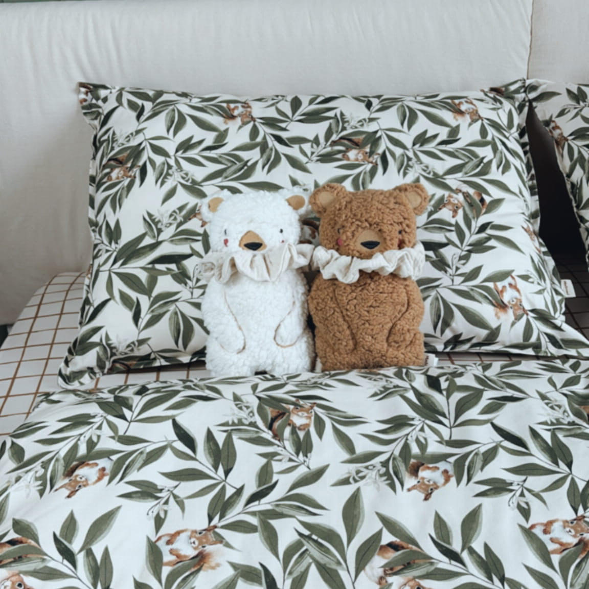 Twee pluche beren met ruches om hun nek zitten op een bed met beddengoed bedrukt met bladeren en eekhoorns. De achtergrond is een groen houten paneel.