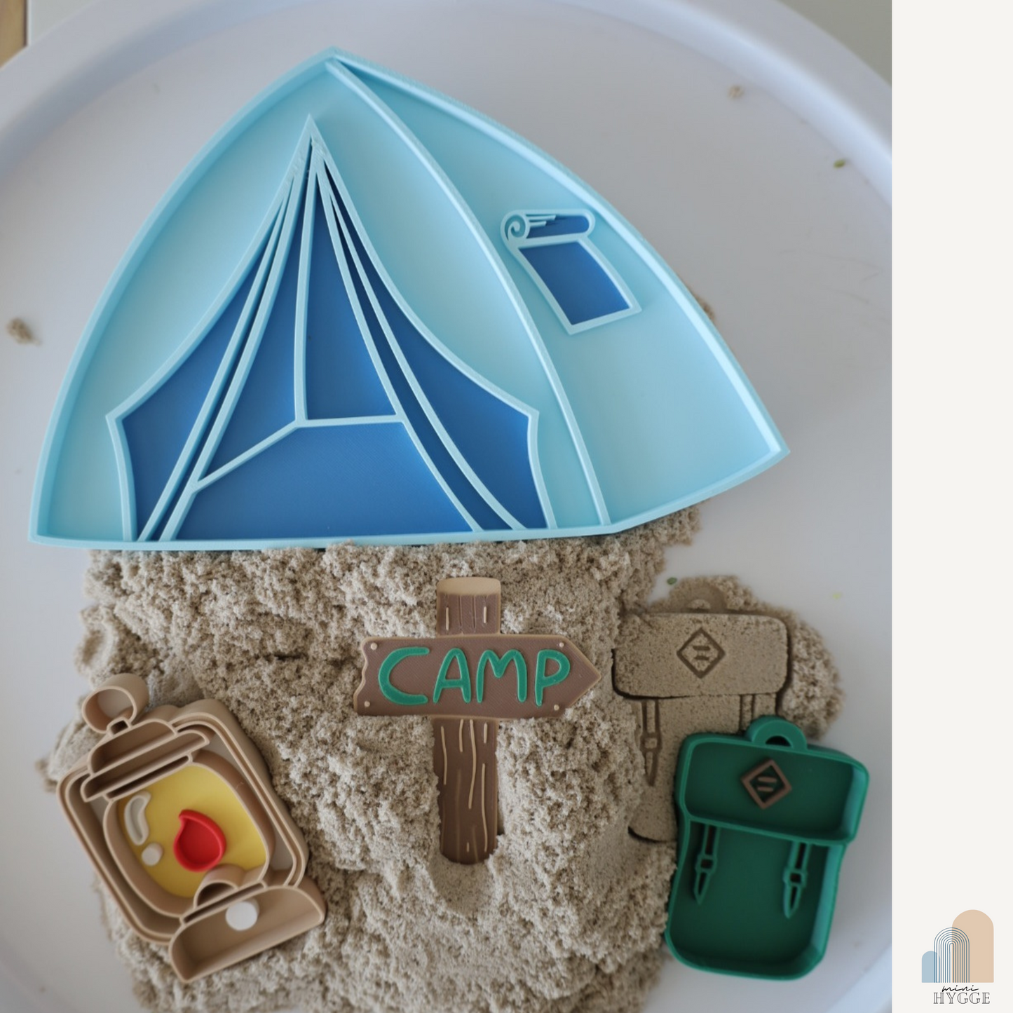Top view of a sensory play setup featuring a blue 3D-printed tent tray placed on kinetic sand, accompanied by three mini trays shaped like a lantern, compass, and backpack.