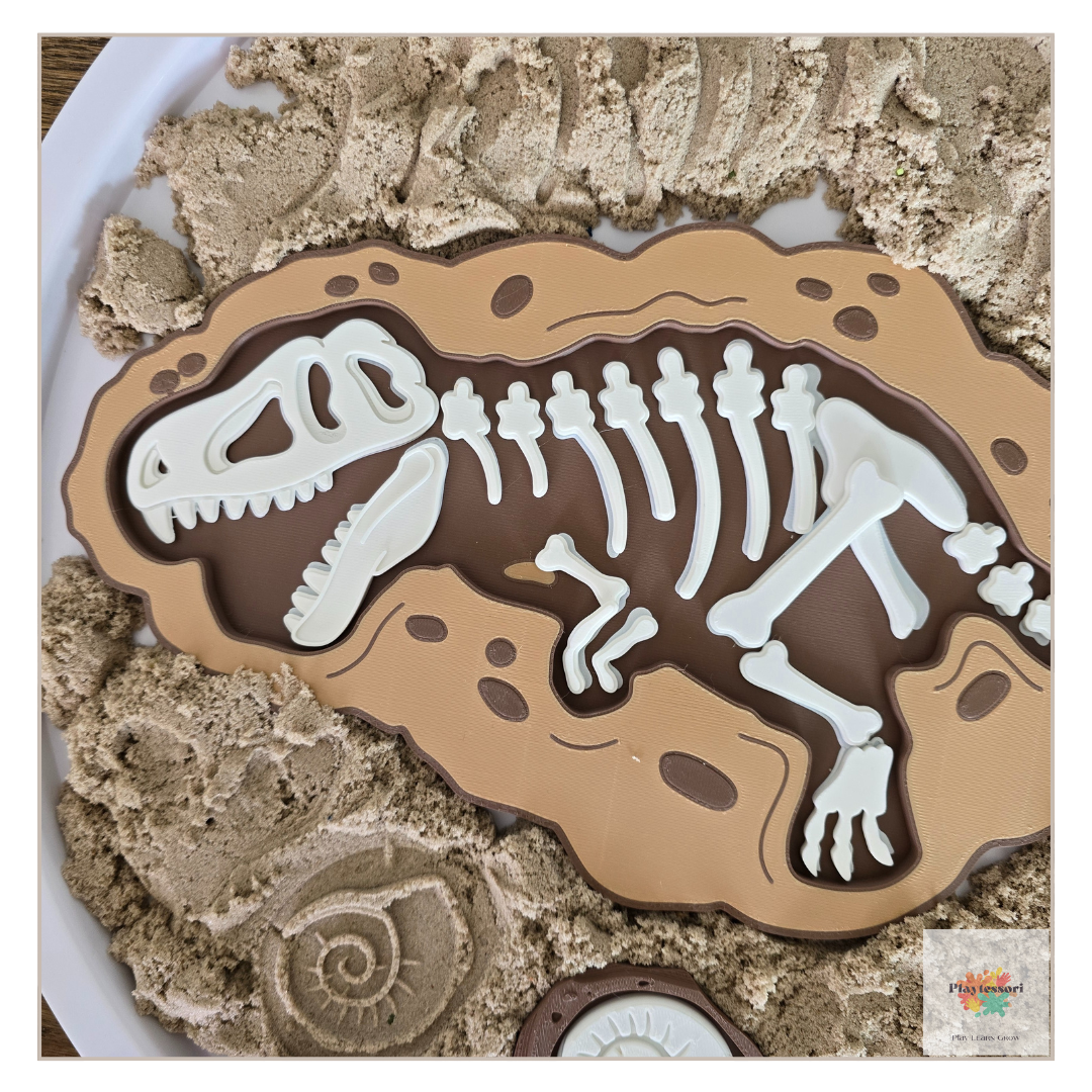 A top-down close-up of a brown, 3D-printed excavation tray with an embossed white T-rex skeleton. The tray sits on beige sensory sand, with a small fossil mold below.