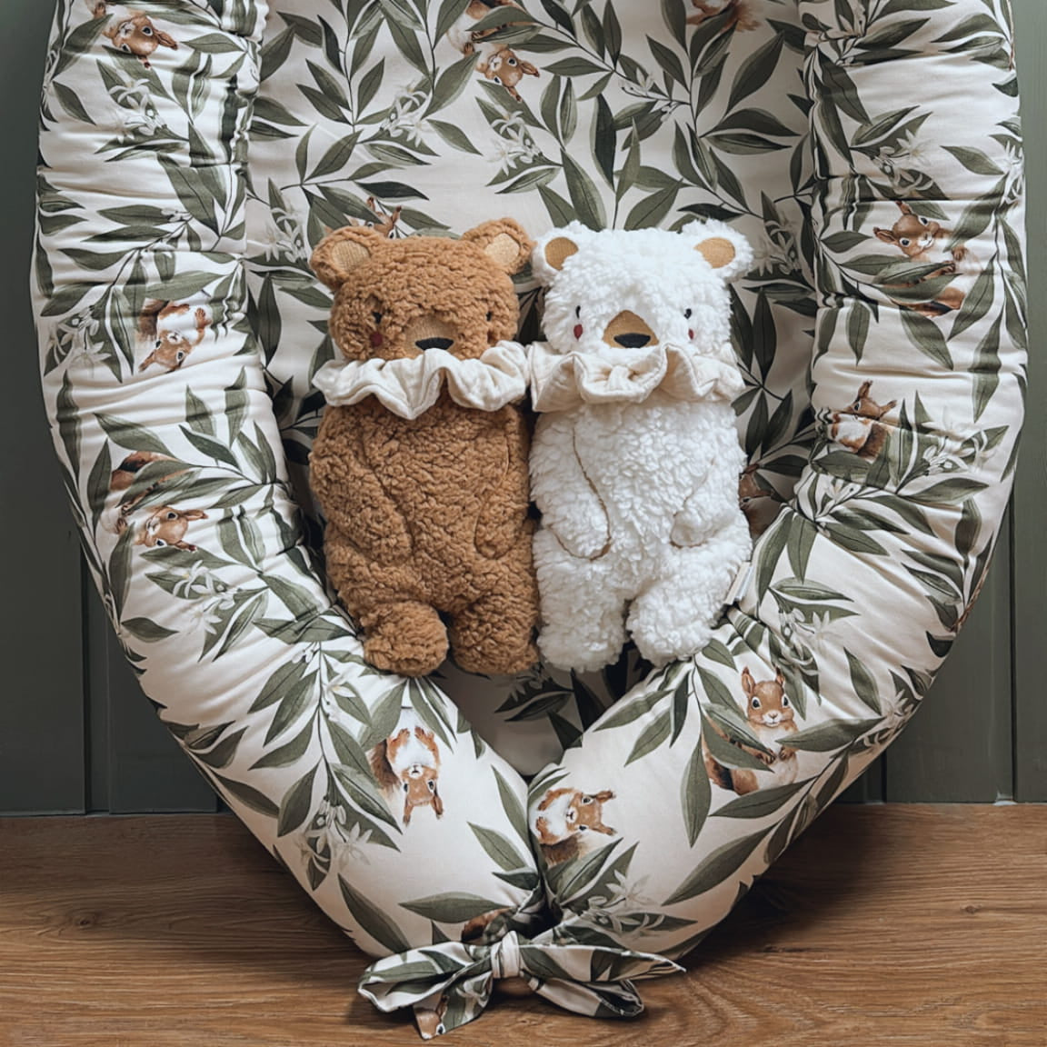 Babynestje met woodlandprint – blaadjes en eekhoorns – met daarin twee knuffelberen, een bruine en een witte, beide met een sierlijke kraag. Nestje staat op een houten vloer tegen een mintgroene houten wand.