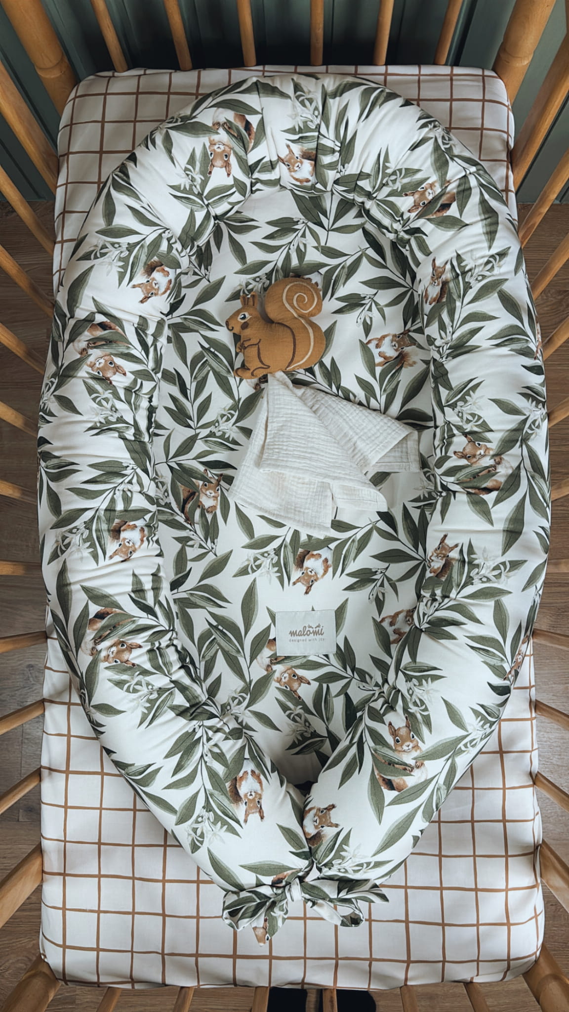 Babynestje met Woodland-patroon (eekhoorns en bladeren) in een houten ledikant, op een geruit hoeslaken. In het nestje liggen een muslin doekje en een zachte bruine eekhoornknuffel.