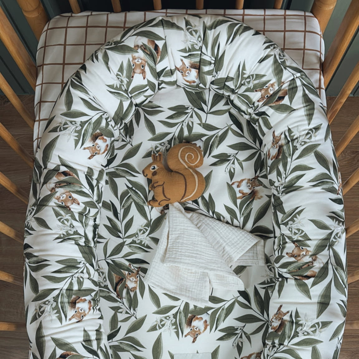 Babynestje met Woodland-patroon (eekhoorns en bladeren) in een houten ledikant, op een geruit hoeslaken. In het nestje liggen een muslin doekje en een zachte bruine eekhoornknuffel.