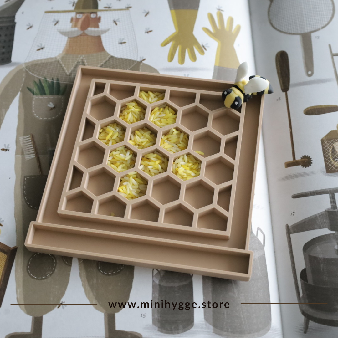 Een bijenkorfvormige sensorische speeltray gevuld met geel en wit gekleurde rijst, vergezeld van een kleine bijenfiguur. De speeltray is gemaakt van bio-gebaseerd PLA en staat op een beige ondergrond met een geillustreerd boek over bijen en imkers op de achtergrond. Onderaan staat de website www.minihygge.store