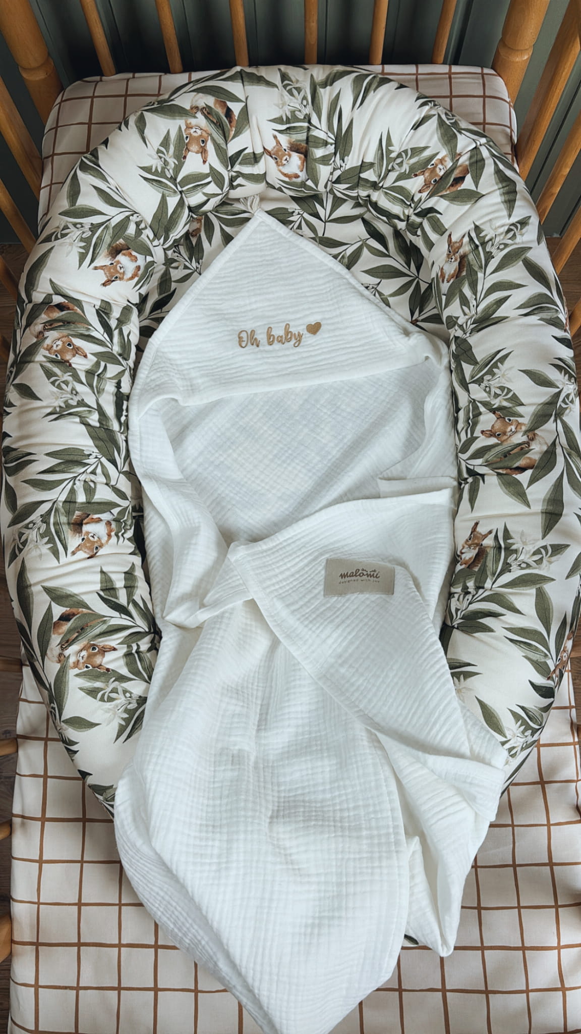 Babynestje met Woodland-patroon in een houten ledikant, met daarin een witte mousseline doek waarop "Oh baby" geborduurd staat in goudkleurige letters.