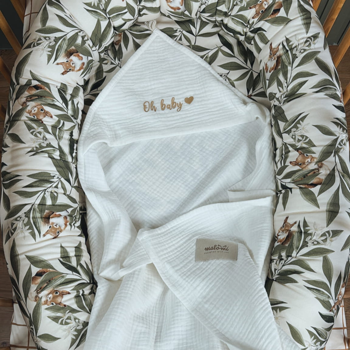 Babynestje met Woodland-patroon in een houten ledikant, met daarin een witte mousseline doek waarop "Oh baby" geborduurd staat in goudkleurige letters.