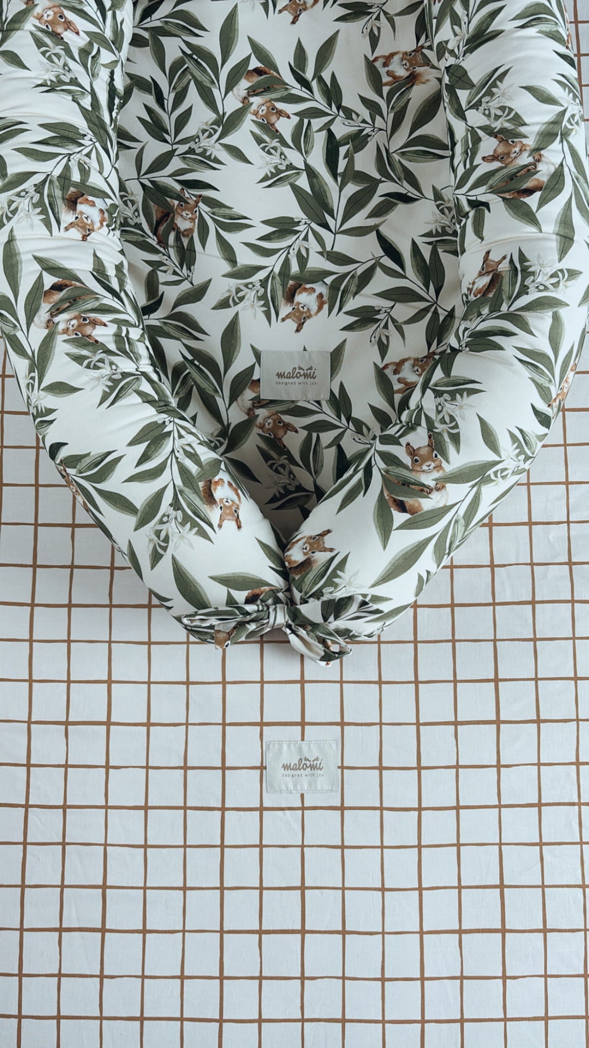 Detail van een babynestje met woodlandprint – blaadjes en eekhoorns – liggend op een geruit hoeslaken met Malomi-label in een babybedje.