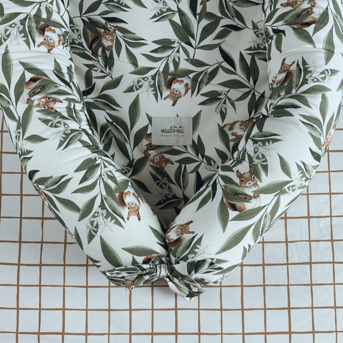 Detail van een babynestje met woodlandprint – blaadjes en eekhoorns – liggend op een geruit hoeslaken met Malomi-label in een babybedje.