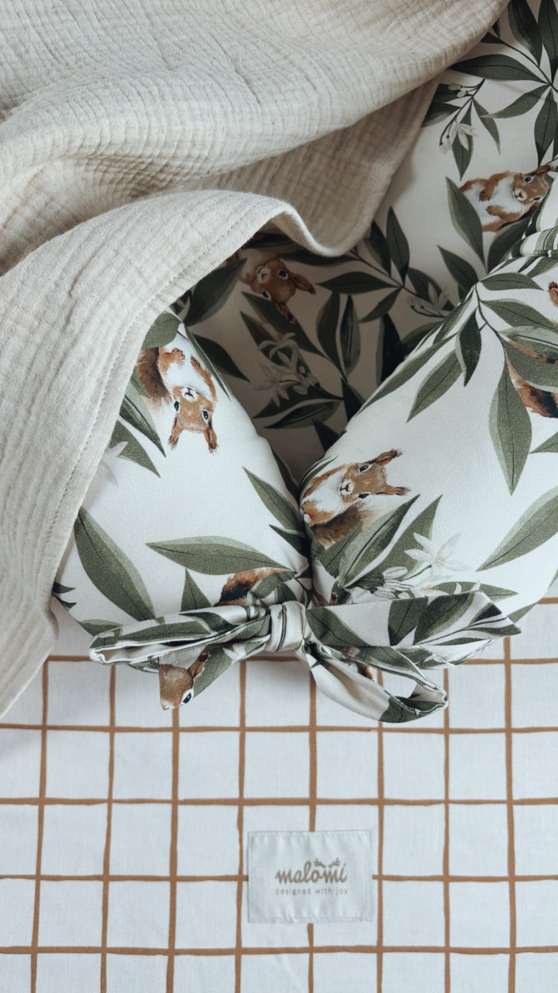 Detailopname van een babynestje met Woodland-patroon, afgewerkt met een strik en bedekt met een beige mousseline doek. Ondergrond: hoeslaken met bruin ruitjespatroon en Malomi-label.