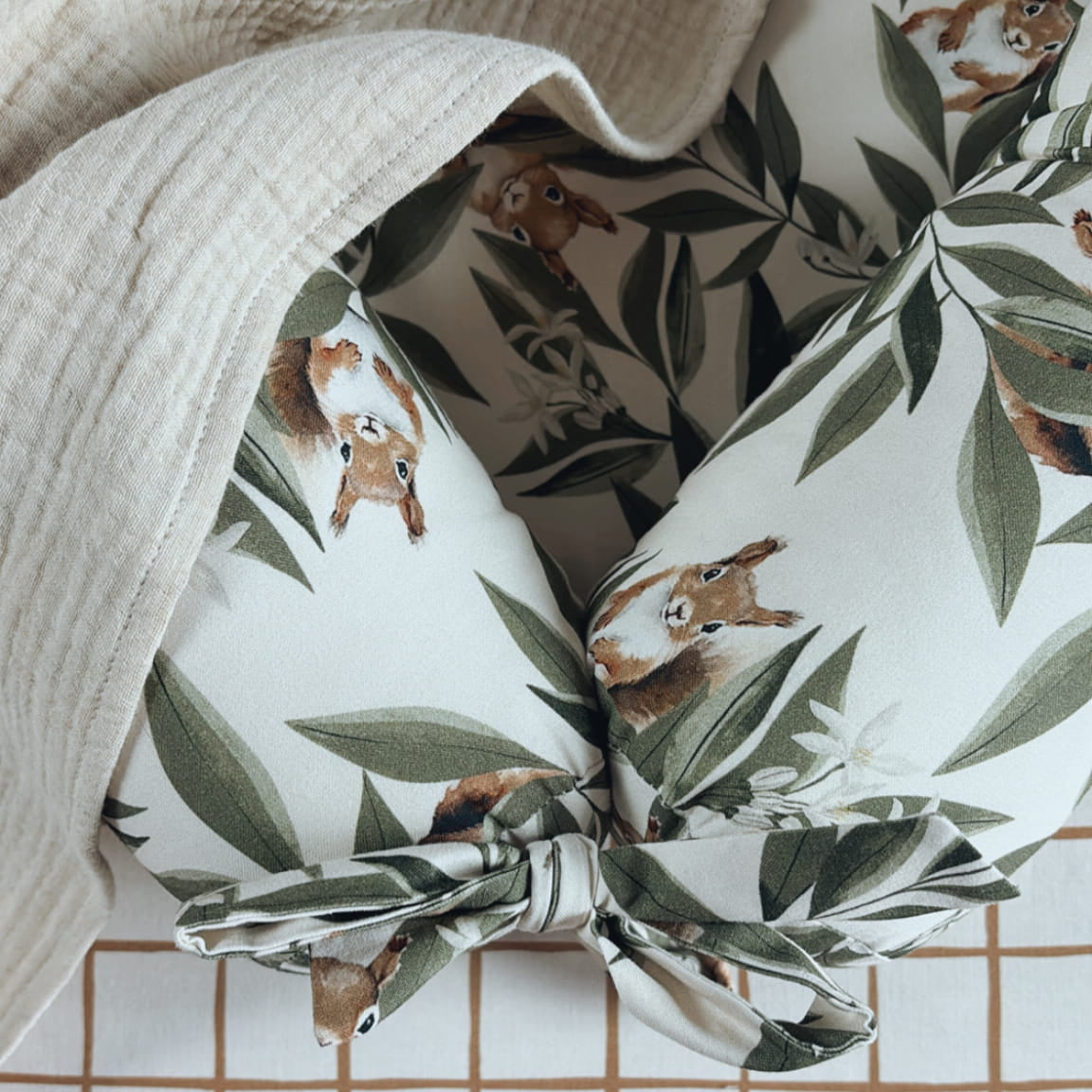 Detailopname van een babynestje met Woodland-patroon, afgewerkt met een strik en bedekt met een beige mousseline doek. Ondergrond: hoeslaken met bruin ruitjespatroon en Malomi-label.