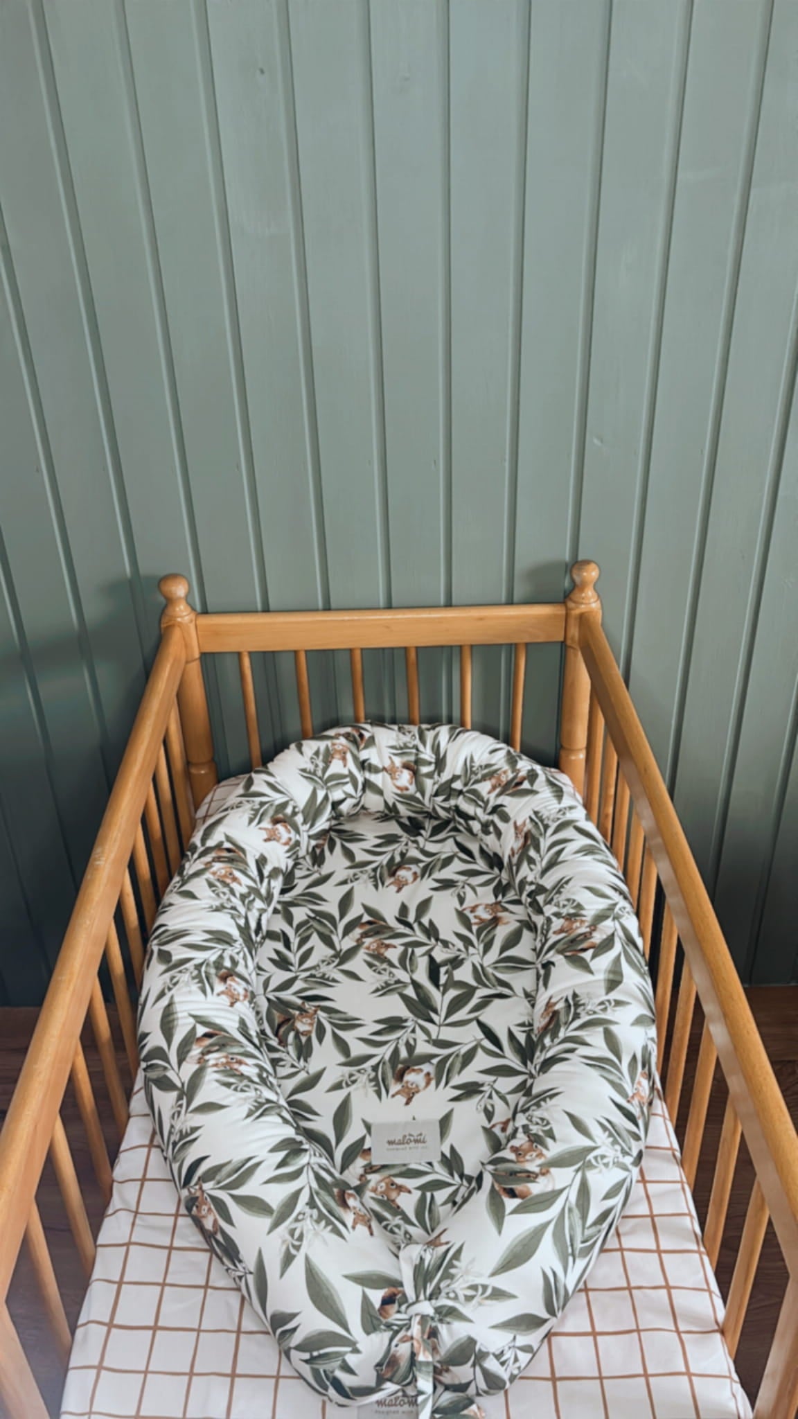 Babynestje met woodlandprint – bladeren en eekhoorntjes – geplaatst in een houten babybedje met geruit hoeslaken. Achtergrond met groene houten wandpanelen.