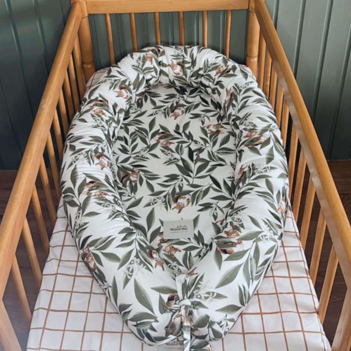 Babynestje met woodlandprint – bladeren en eekhoorntjes – geplaatst in een houten babybedje met geruit hoeslaken. Achtergrond met groene houten wandpanelen.