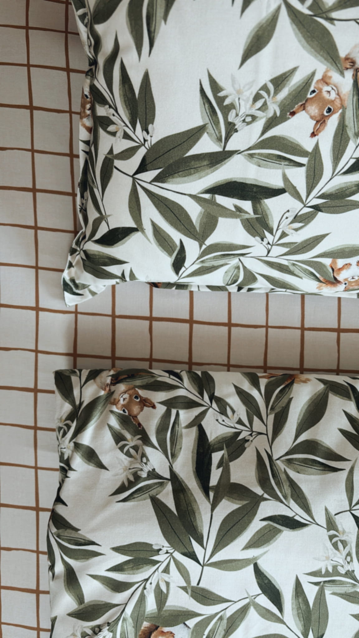Detail van beddengoed met een patroon van groene bladeren en kleine eekhoorns op een witte achtergrond. Op de achtergrond is een beige hoeslaken met een bruin ruitjespatroon te zien.