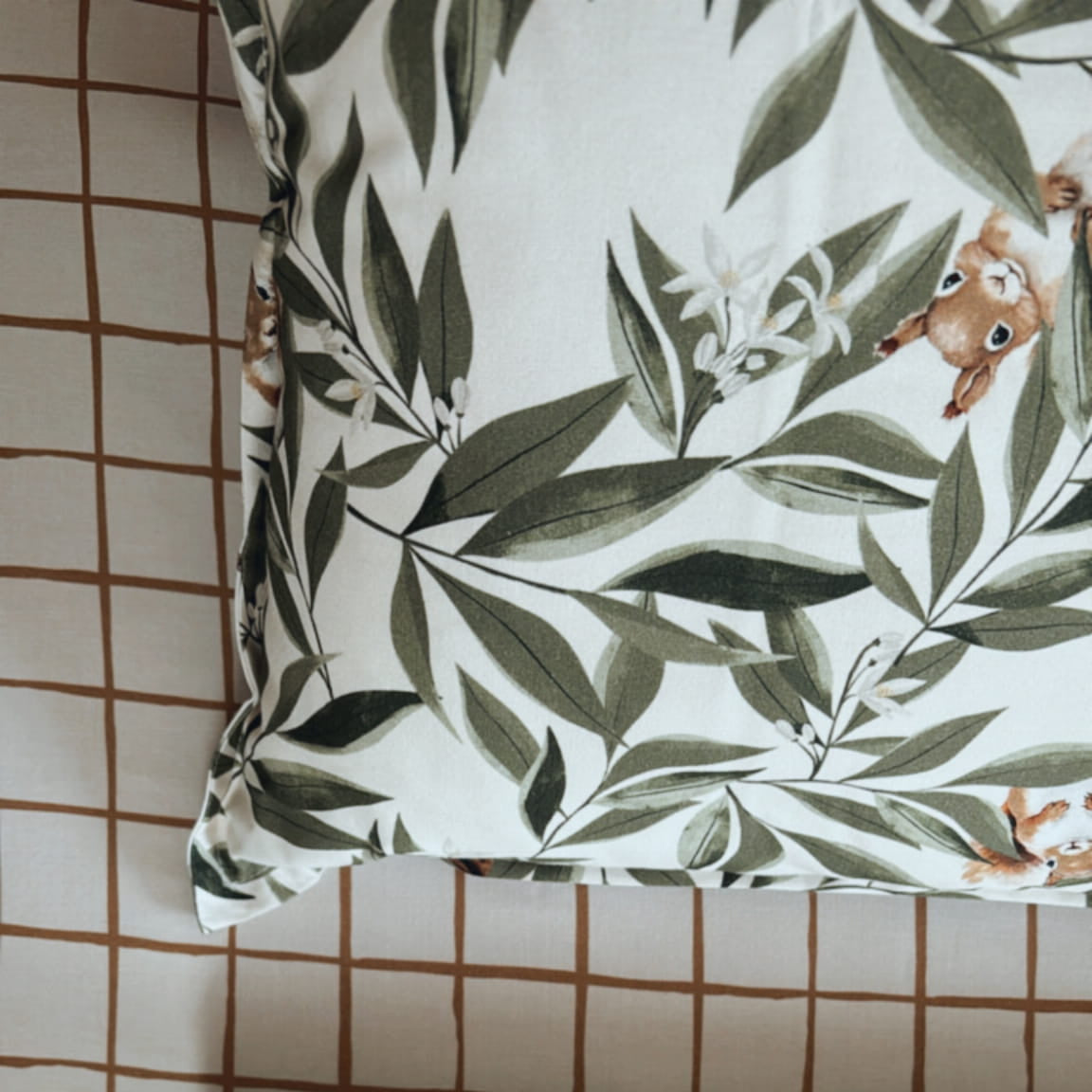 Detail van beddengoed met een patroon van groene bladeren en kleine eekhoorns op een witte achtergrond. Op de achtergrond is een beige hoeslaken met een bruin ruitjespatroon te zien.