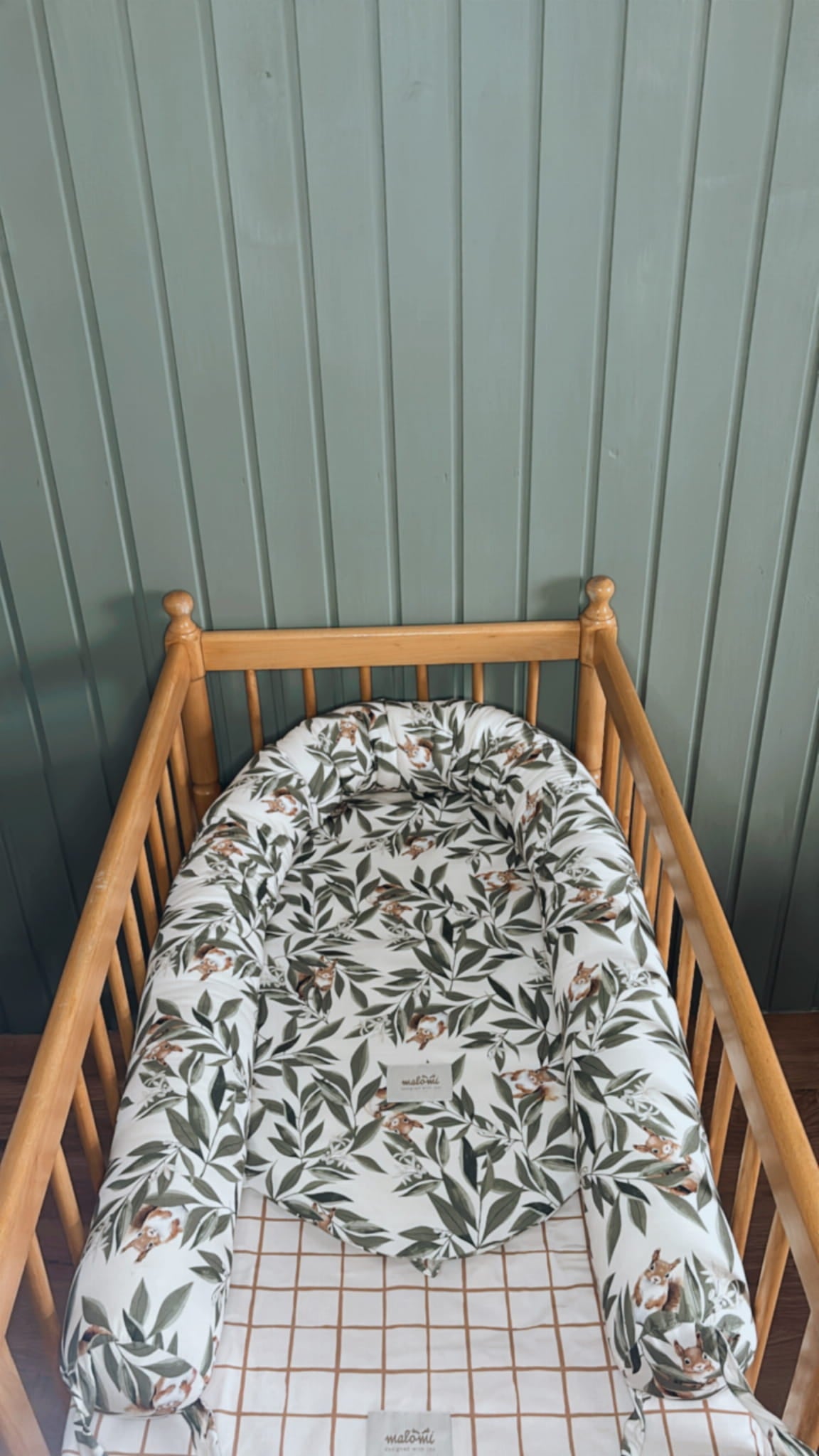 Babynestje met Woodland-patroon in een houten ledikantje, geplaatst op een hoeslaken met bruin ruitjesmotief. Het nestje toont een schattig ontwerp met blaadjes en eekhoorntjes.