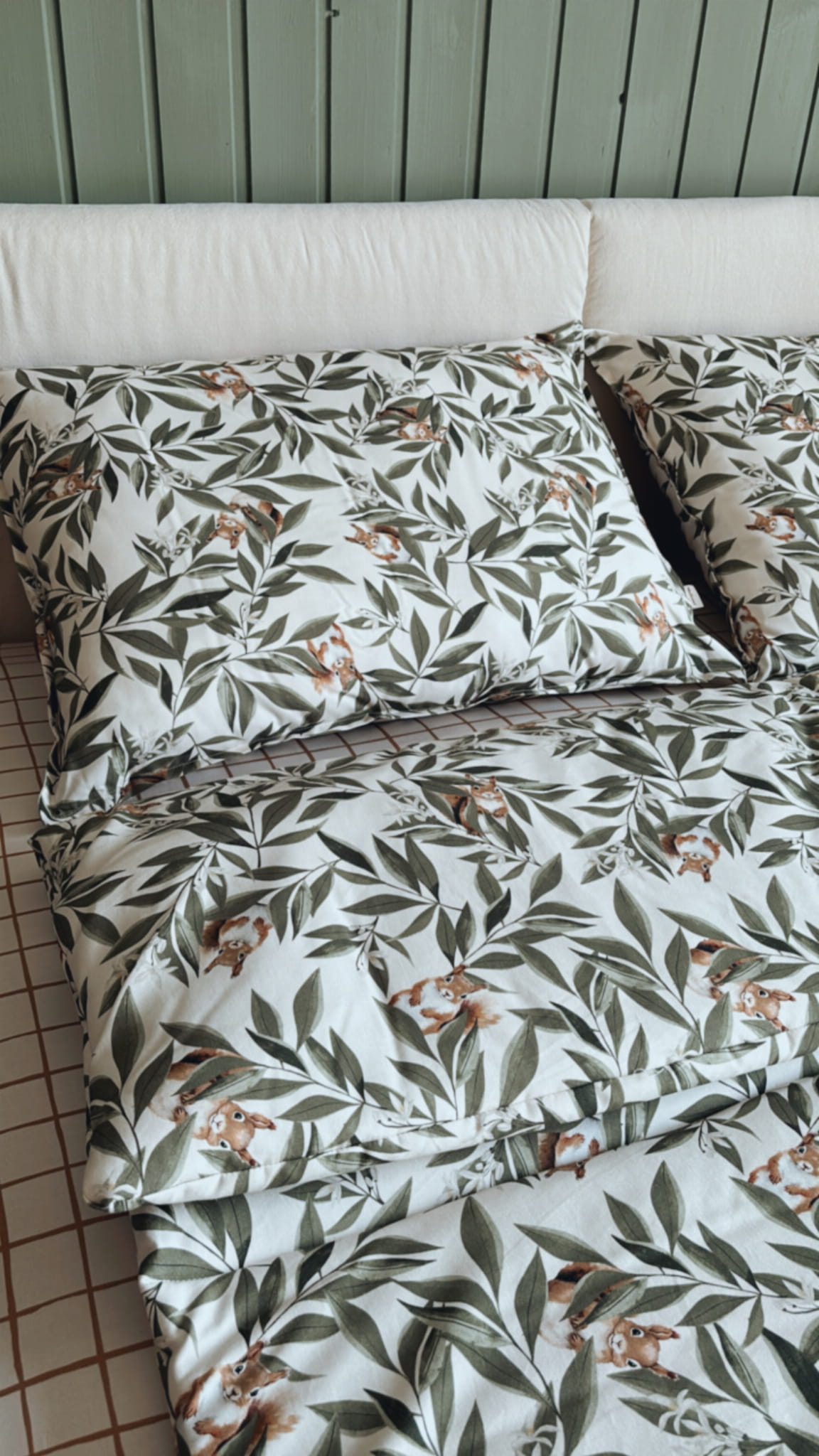 Opgemaakt bed met kussens en dekbedovertrek met een patroon van groene bladeren en speelse eekhoorns. De achtergrond bestaat uit een groen houten hoofdbord en een hoeslaken met een bruin ruitjesmotief.