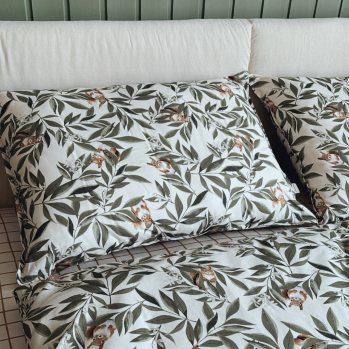 Opgemaakt bed met kussens en dekbedovertrek met een patroon van groene bladeren en speelse eekhoorns. De achtergrond bestaat uit een groen houten hoofdbord en een hoeslaken met een bruin ruitjesmotief.