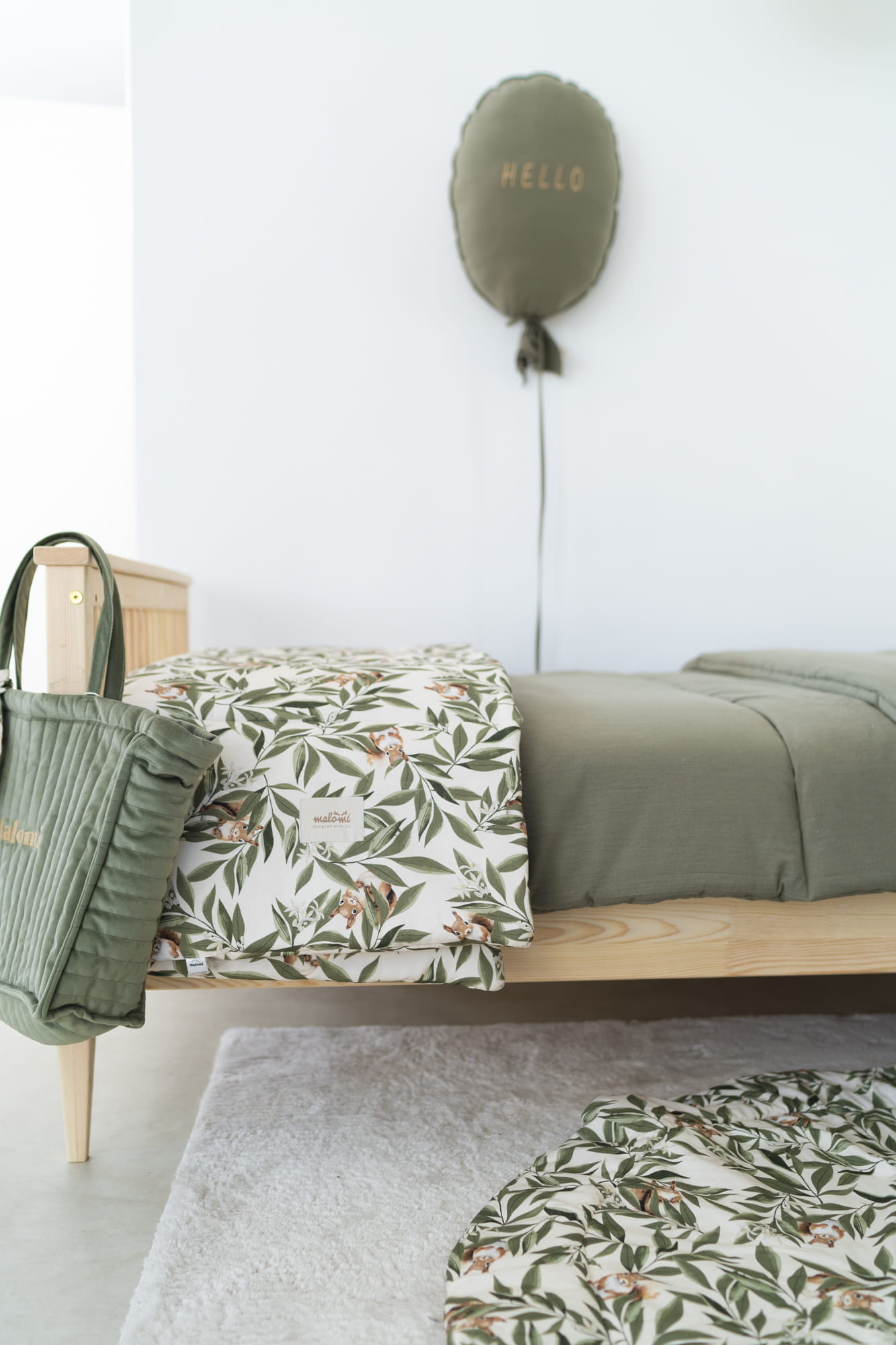 Kinderkamer met een houten bed waarop een dekbedovertrek met eekhoorn- en bladerenprint ligt. Naast het bed hangt een groene gewatteerde tas en op de muur een ballonvormig kussen met het woord 'HELLO'. Op de vloer ligt een wit vloerkleed en een bijpassend rond speelkleed.