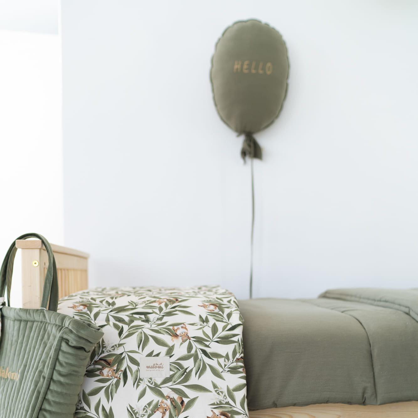 Kinderkamer met een houten bed waarop een dekbedovertrek met eekhoorn- en bladerenprint ligt. Naast het bed hangt een groene gewatteerde tas en op de muur een ballonvormig kussen met het woord 'HELLO'. Op de vloer ligt een wit vloerkleed en een bijpassend rond speelkleed.