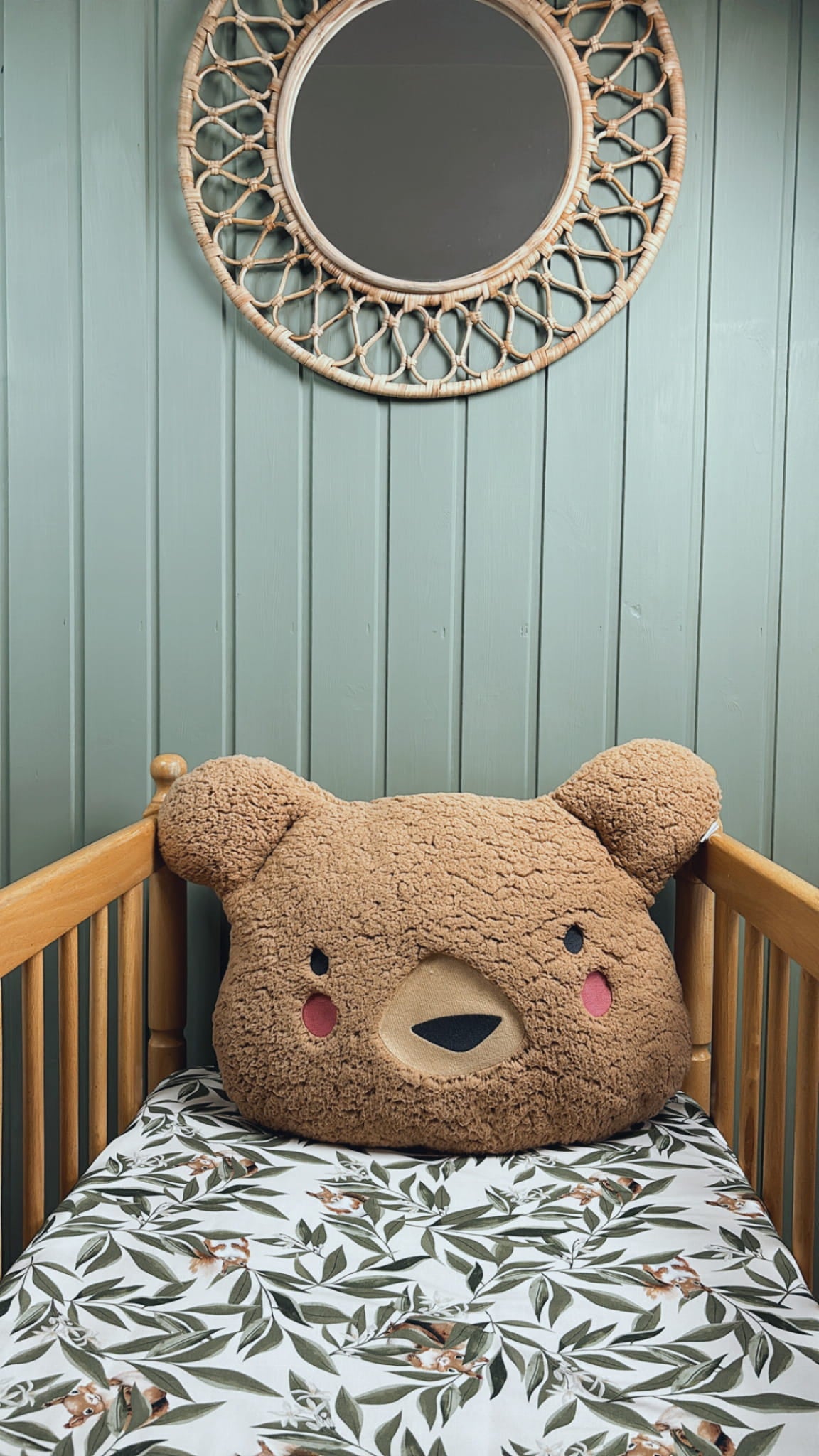 Kinderbedje met woodland hoeslaken met bladeren en eekhoorns, versierd met een groot pluche berenkussen. Aan de muur hangt een ronde rotan spiegel boven een groene houten wand.