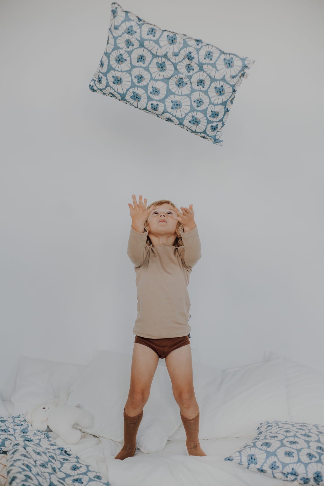 Kind gooit kussen omhoog op een wit bed – patroon denim leeuwen zichtbaar.
