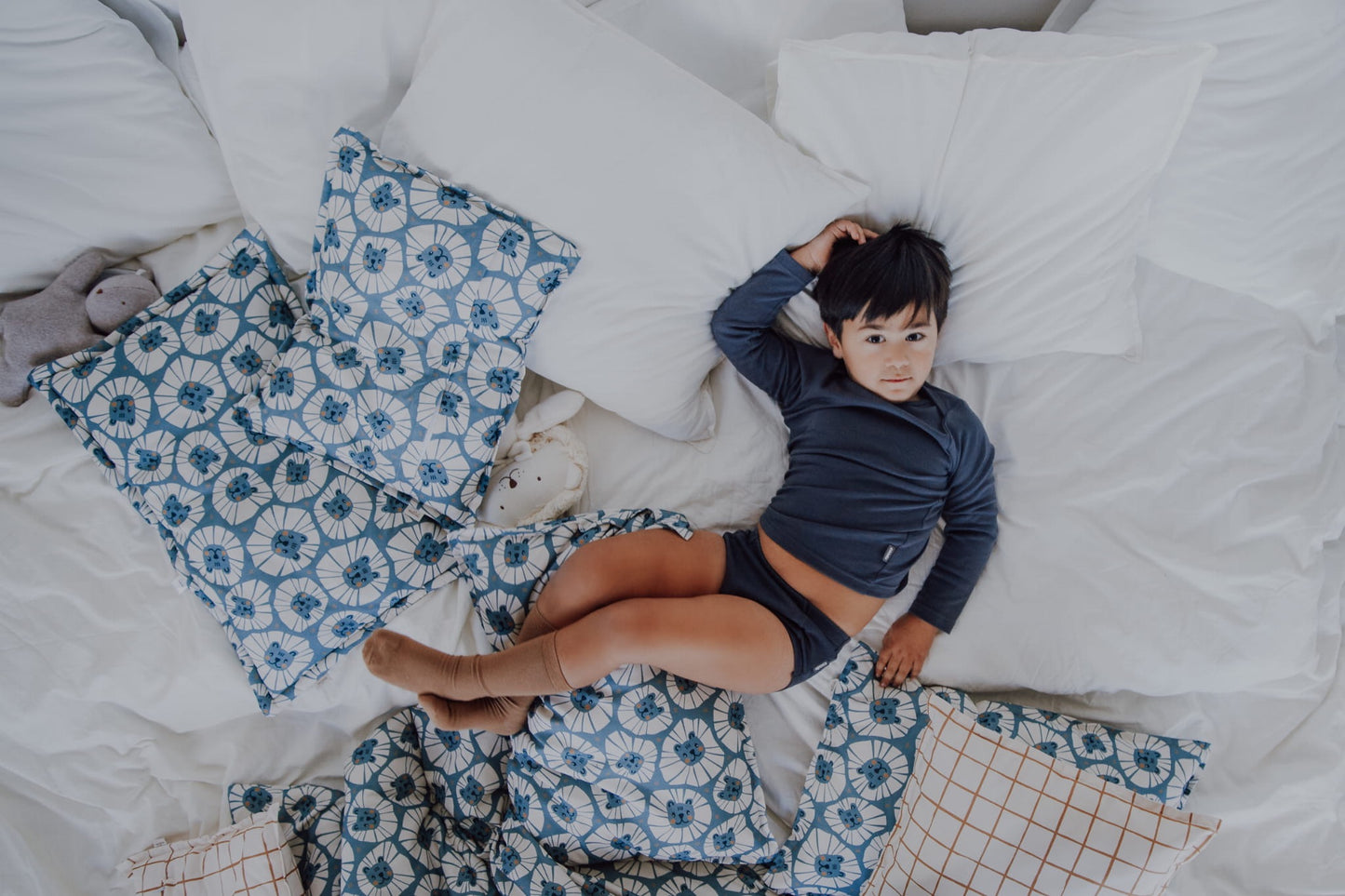 Jongen ligt ontspannen op wit beddengoed met denim leeuwen kussen en deken.