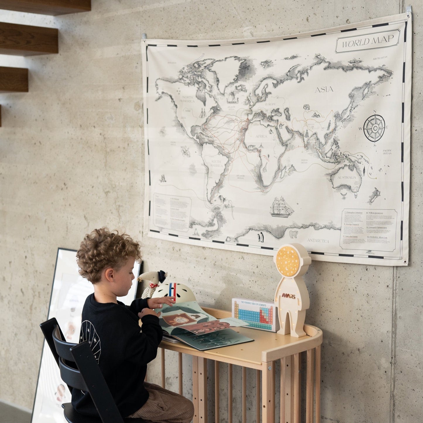 A child sits at a wooden desk reading a book, with a cotton world map 'Travellers and Their Trails' displayed on the wall. The scene is complemented by a modern black chair, colorful stacked cushions, and educational materials, creating a cozy and inspiring learning space