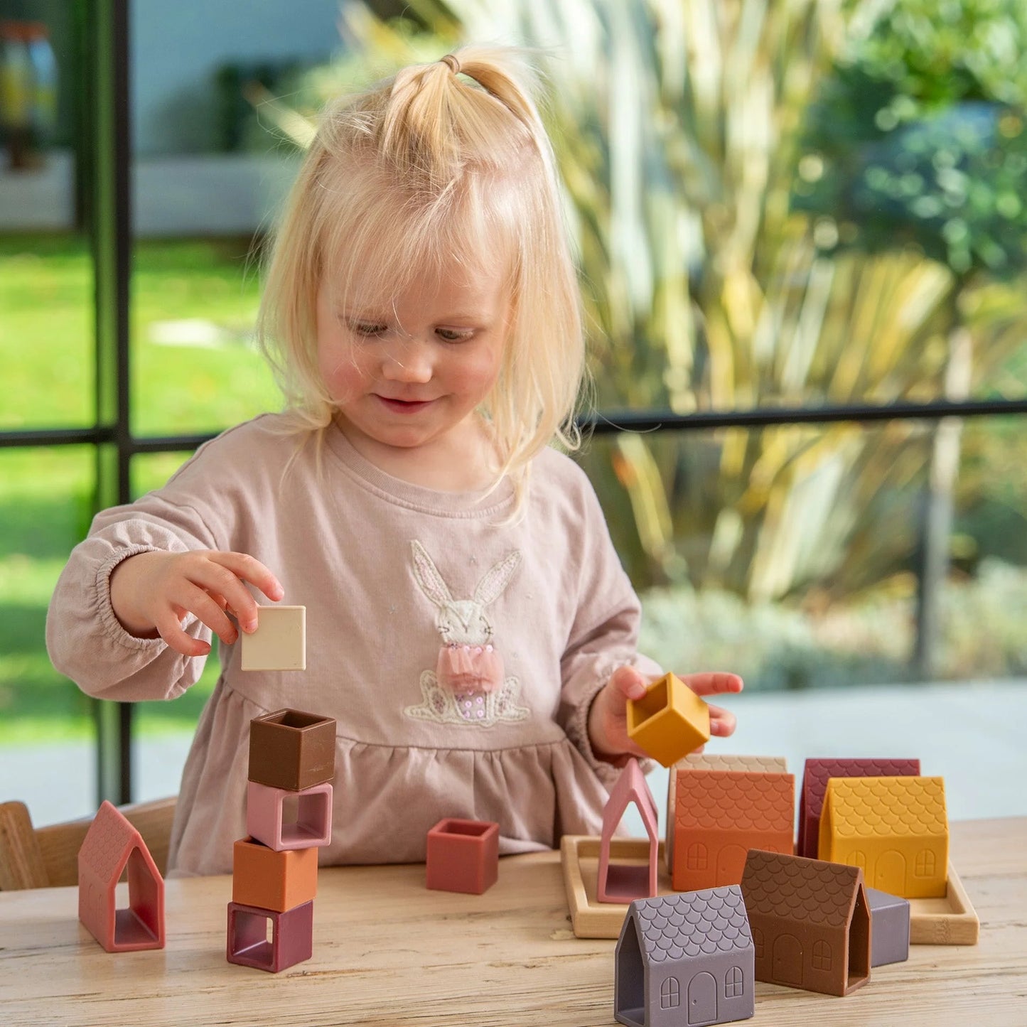 A smiling young child with blonde hair stacking colorful silicone cubes while playing with toy houses on a wooden table