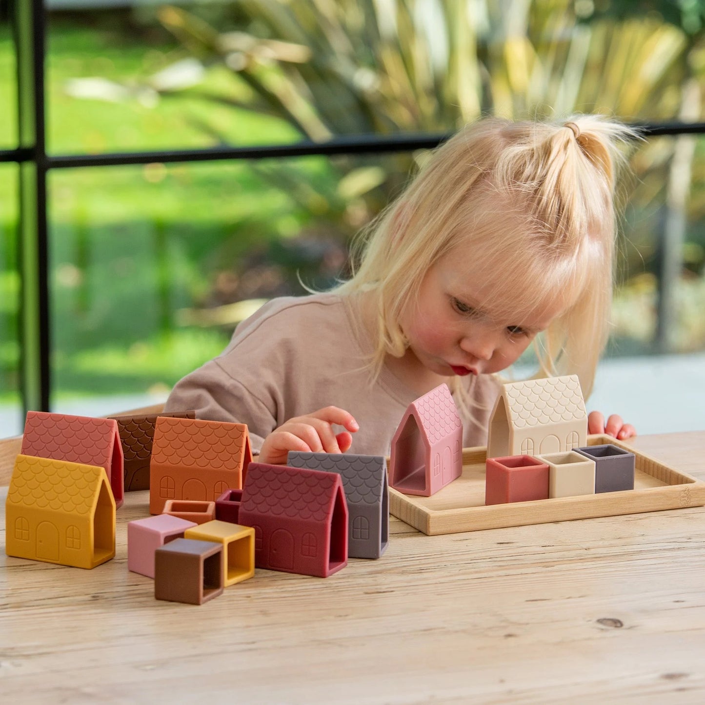 A young child with blonde hair playing intently with colorful silicone toy houses and cubes on a wooden table, holding a yellow house