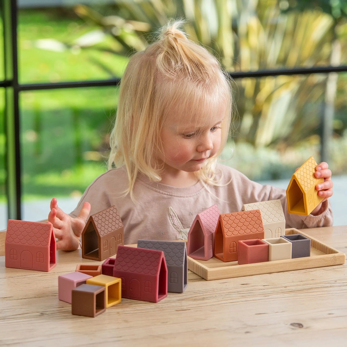 A young child with blonde hair looking down and playing with silicone toy houses and cubes on a wooden table, with a bamboo tray nearby