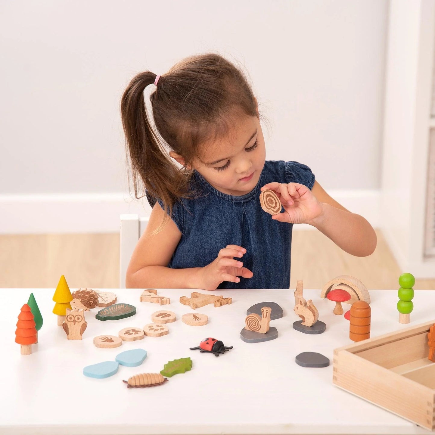 Klein meisje speelt aan tafel met houten dieren uit de TickiT Bospadenset, omringd door natuuraccessoires en boomschijfjes.