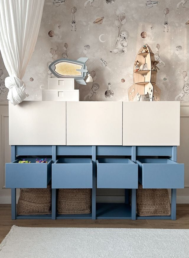 Een opgeruimde Tweens commode met gesloten witte deuren en vier open lades in muted blauw, perfect voor manden. Het meubel staat in een gezellige kinderkamer met leuk ruimte-thema behang en een wit hemelgordijn.