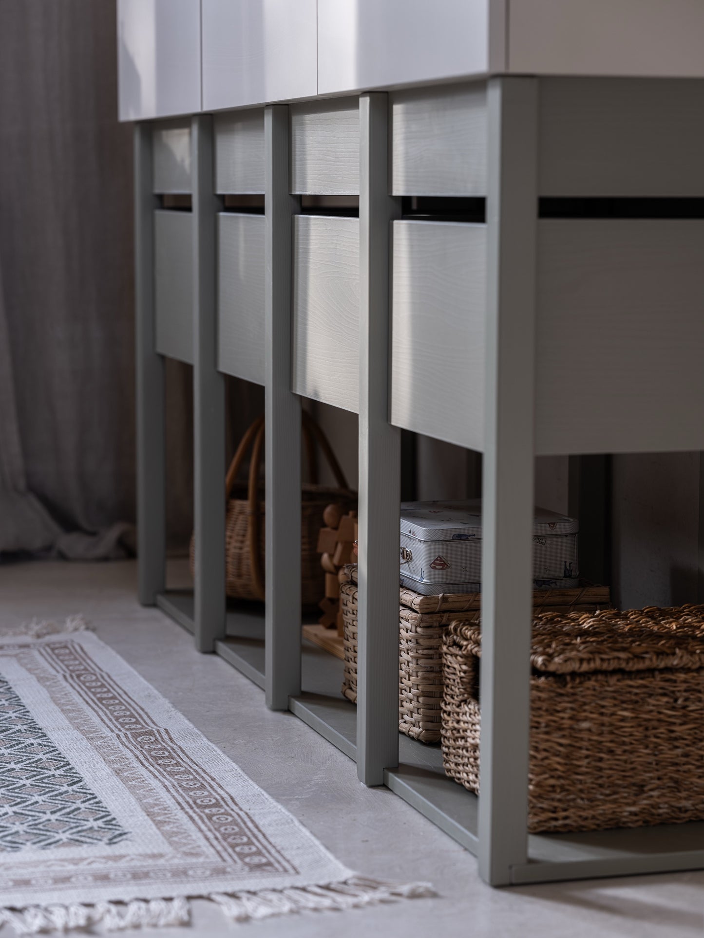 Een close-up van de onderste sectie van de Tweens commode in eucalyptusgroen, met lades en open vakken gevuld met rieten manden en speelgoed. De textuur van het meubel en de gezellige vloerbedekking zijn goed zichtbaar.