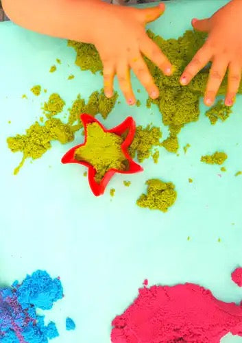 Kind speelt met felgroen dynamisch zand op een blauwe tafel. Het zand wordt gevormd met een rode stervormige mal. Ook blauw en roze zand zijn zichtbaar op de achtergrond.