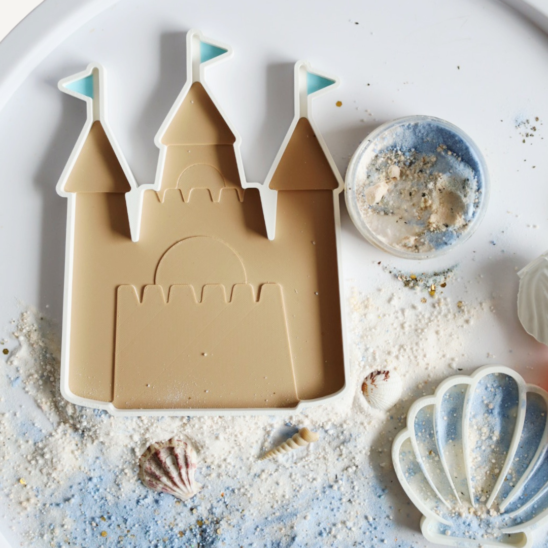 Een strand- en kasteelvormige uitsteker set, inclusief een potje zandkleurige en blauwe speelklei, versierd met glitter en kleine schelpjes.