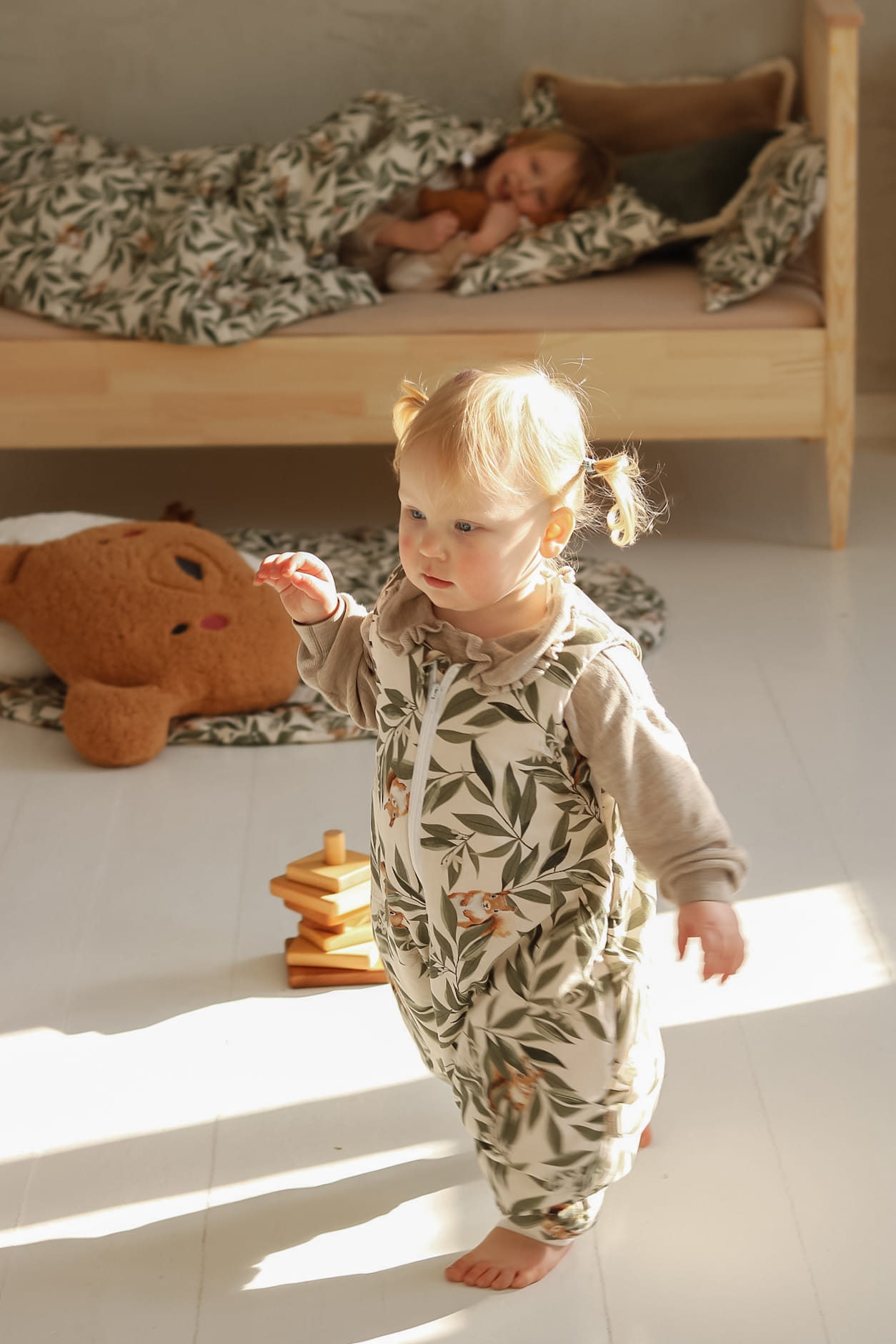 Peuter in een babyslaapzak met woodland print speelt op een zonnige vloer. Op de achtergrond ligt een ouder kind onder bijpassend beddengoed op een houten peuterbed.