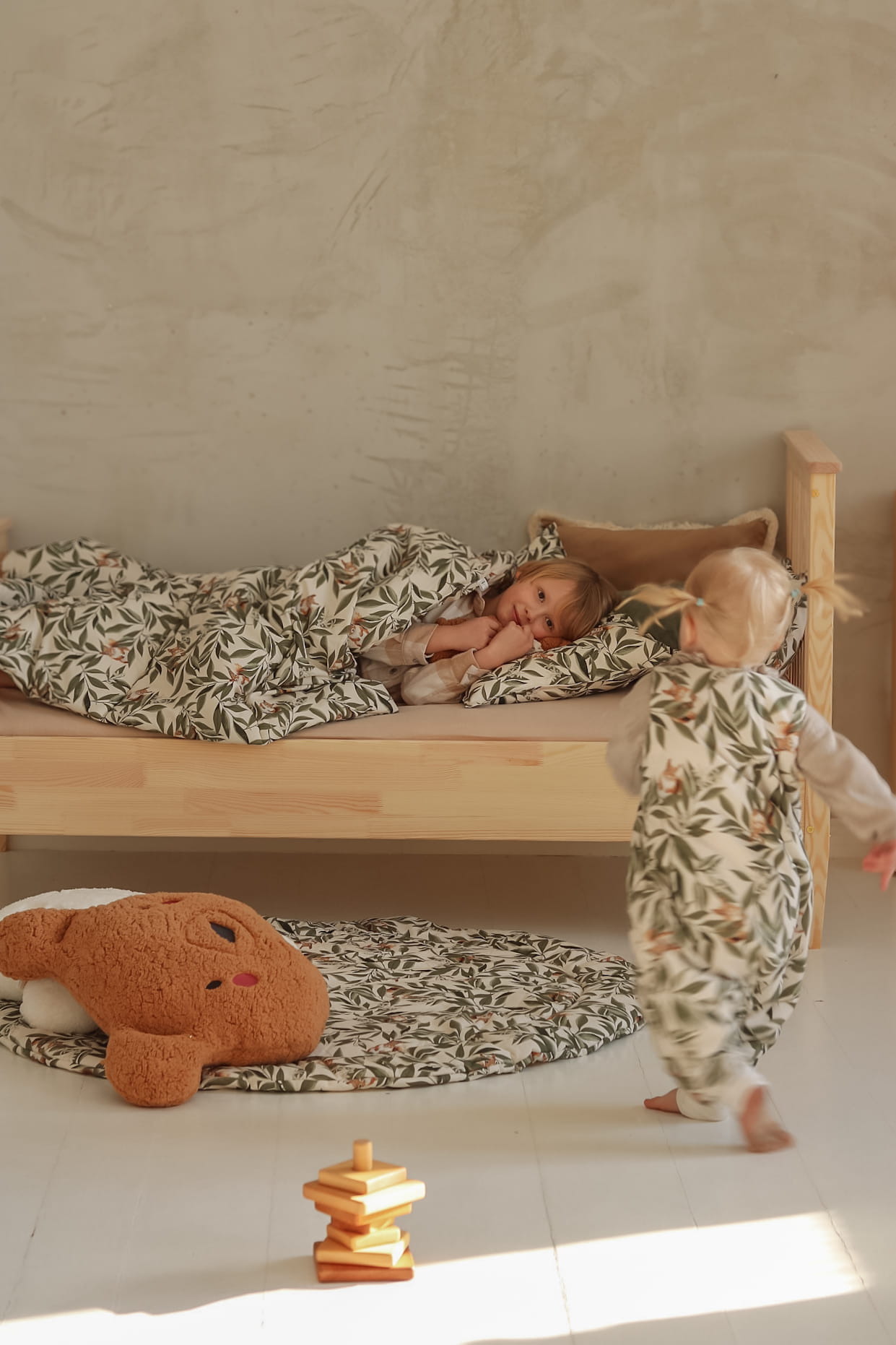Peuter in een slaapzak met woodland print loopt naar een houten bed waarin een ouder kind ligt onder een dekbed met hetzelfde motief. Op de grond liggen een ronde speelmat en een knuffelbeer.