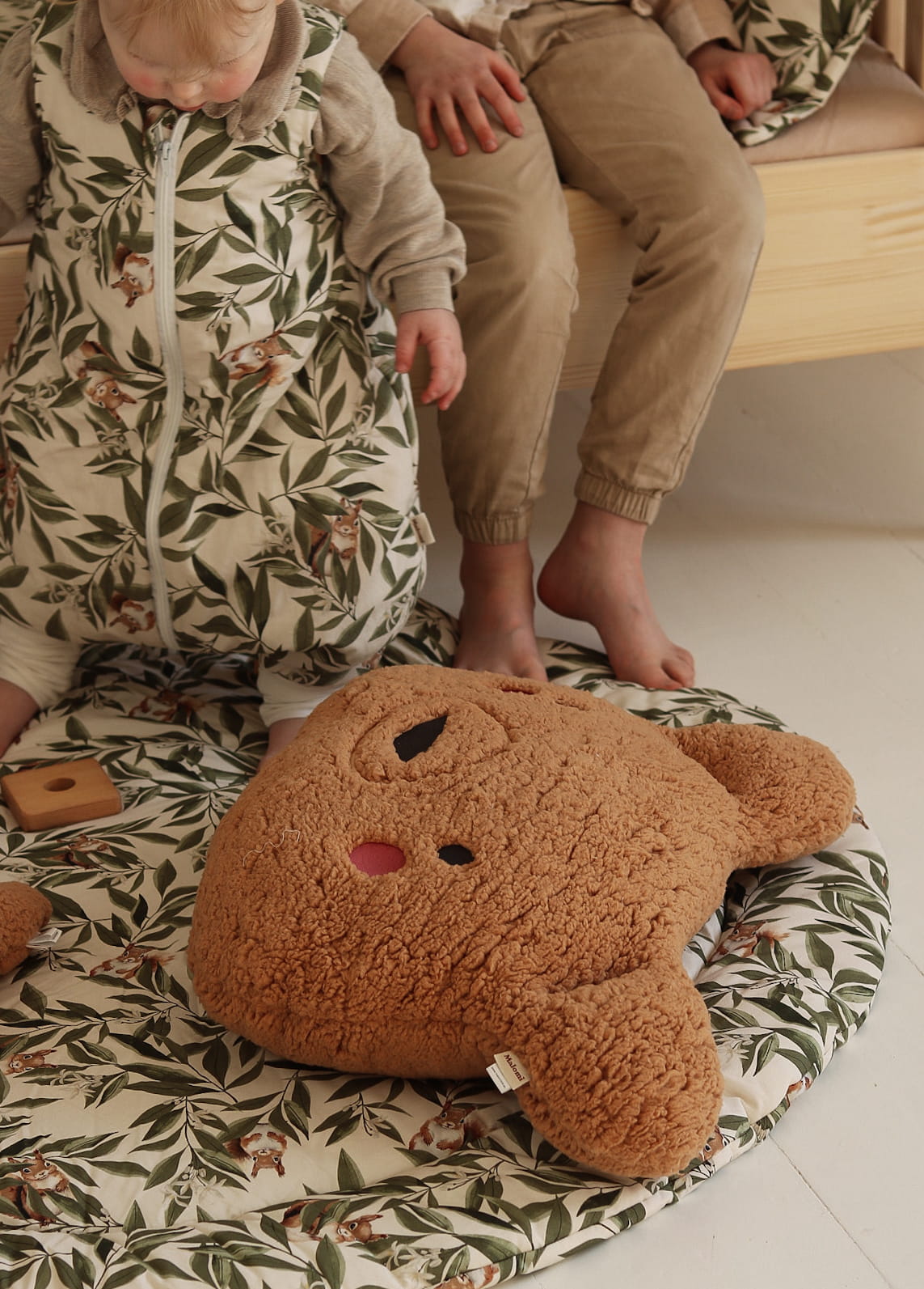 Close-up van een peuter in een slaapzak met woodland print, zittend op een ronde speelmat met hetzelfde motief. Naast het kindje zit een ouder kind op een houten bed. Op de mat ligt een grote pluchen berenknuffel.
