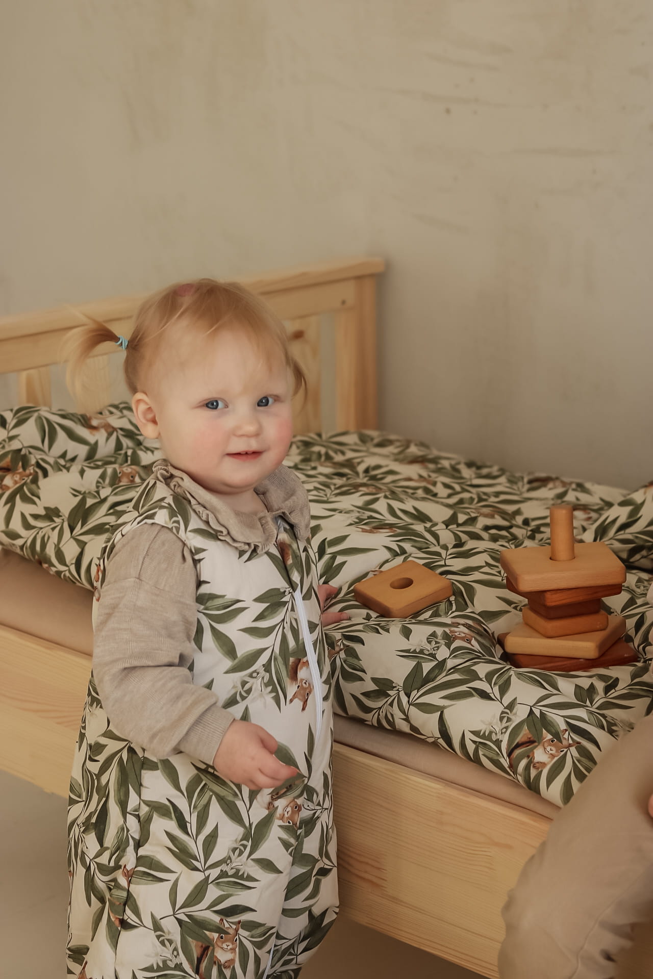 Lachende peuter in een slaapzak met woodland print staat naast een houten bed met bijpassend beddengoed. Op het bed liggen houten stapelblokken.