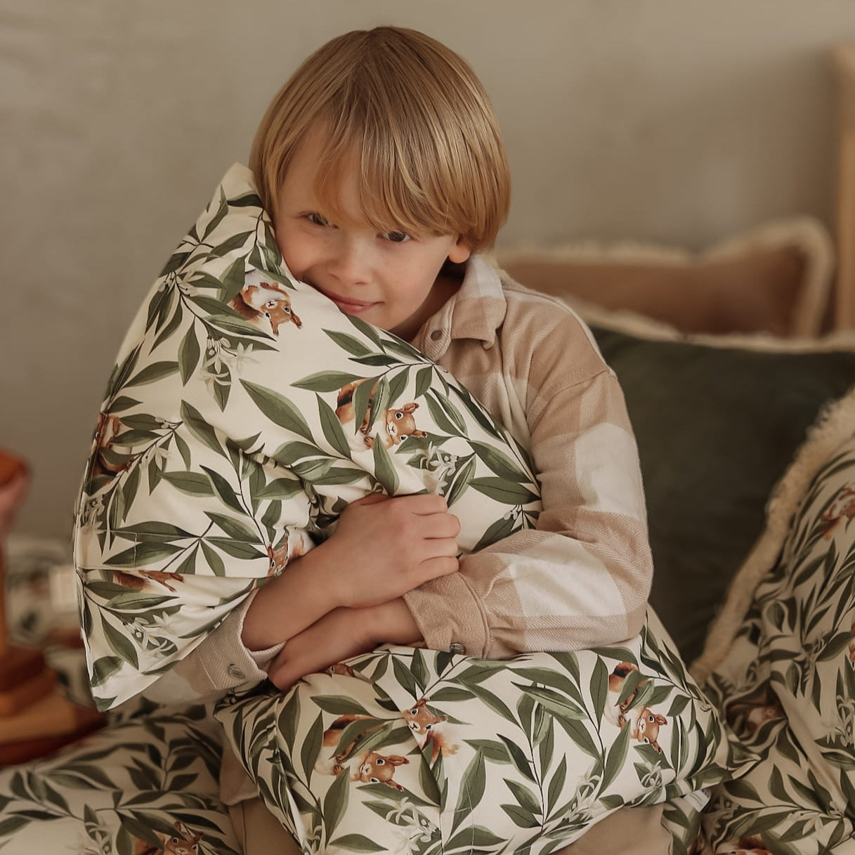 Lachend kind zit op een houten bed en knuffelt een kussen met bladeren- en eekhoornprint. De sfeer is warm en huiselijk, met natuurlijke kleuren en zachte materialen op de achtergrond.
