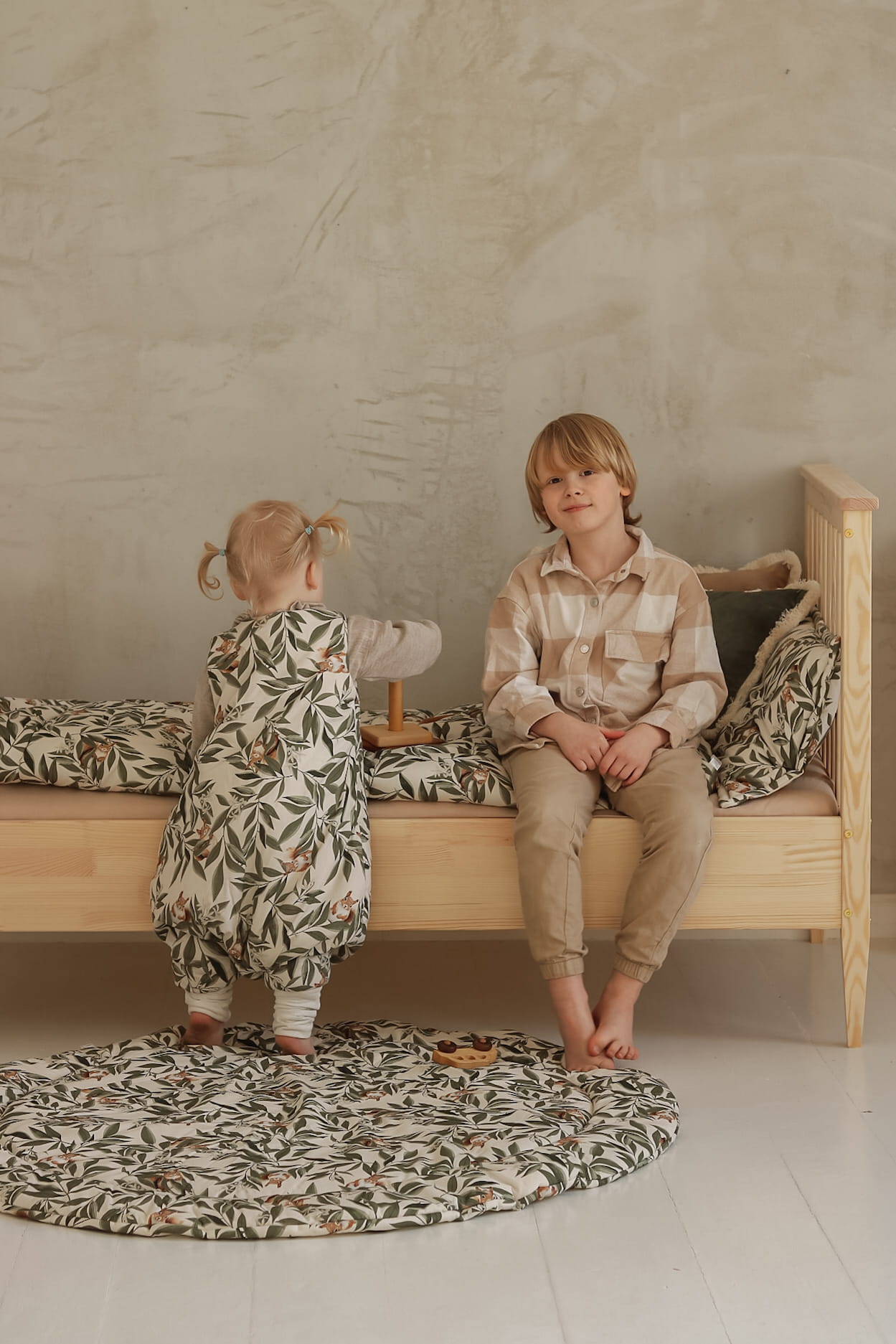 Peuter in een woodland slaapzak speelt met houten speelgoed op een bed met bijpassend beddengoed. Een ouder kind zit glimlachend naast haar op het houten bed, met een ronde speelmat op de vloer.