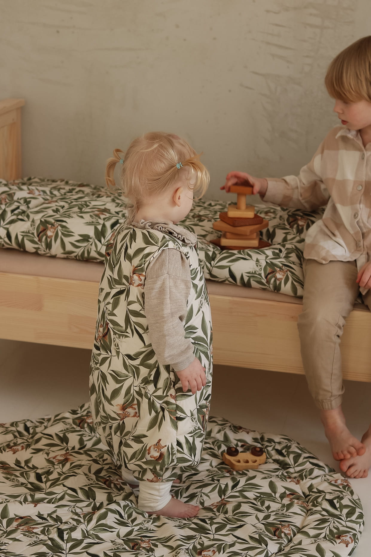 Peuter in een slaapzak met woodland print staat op een bijpassende ronde speelmat en kijkt naar een ouder kind dat houten speelgoed op een bed met hetzelfde motief stapelt.