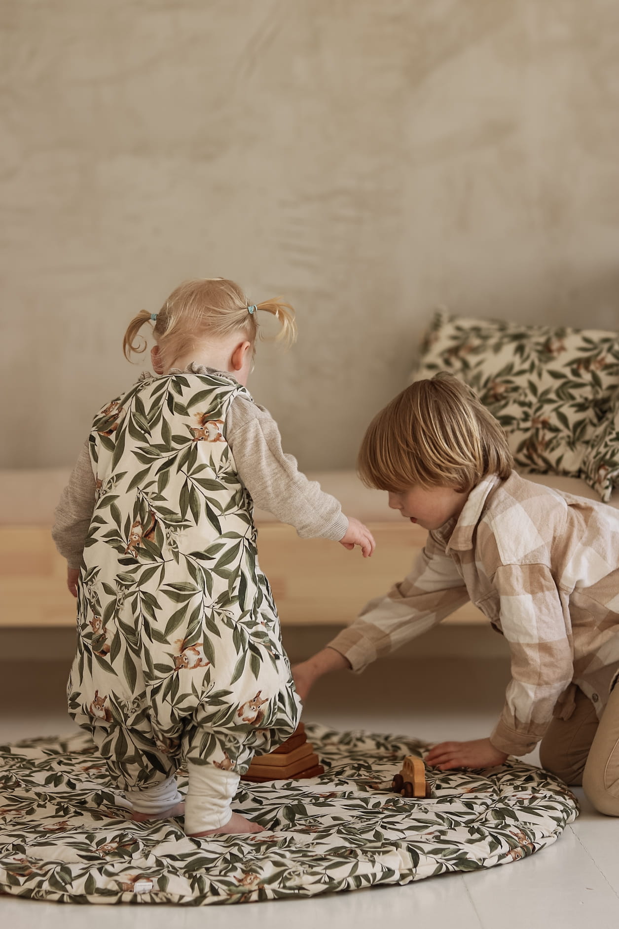 Peuter in een slaapzak met woodland print speelt op een ronde speelmat met een ouder kind. Op de achtergrond ligt een bed met bijpassende kussens en dekens.