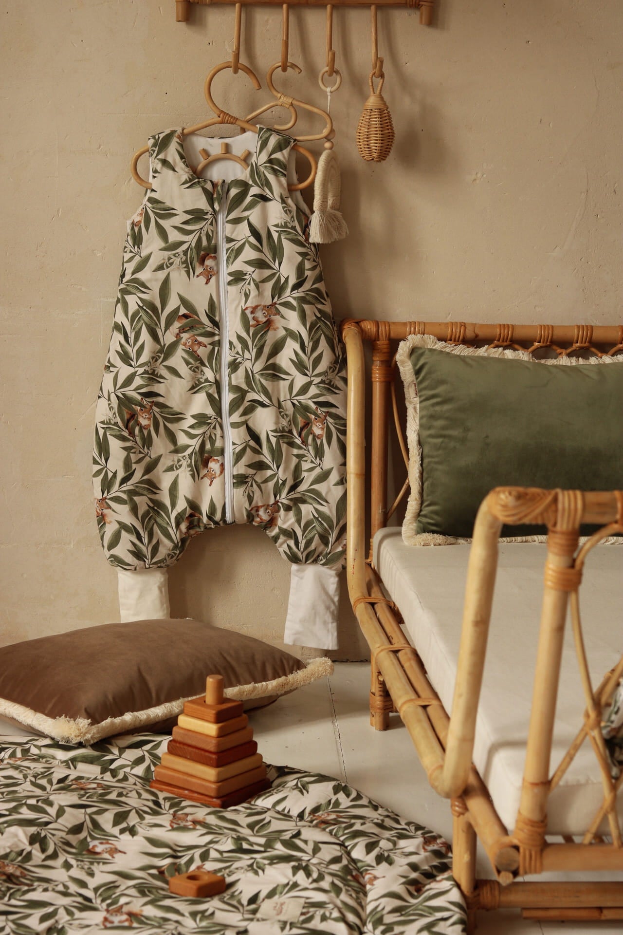 Slaapzak met woodland print hangt aan een rotan kapstok in een kinderkamer met natuurlijke tinten. Op de vloer ligt een speelmat met bijpassende print, samen met een stapel houten speelgoed en een sierkussen.