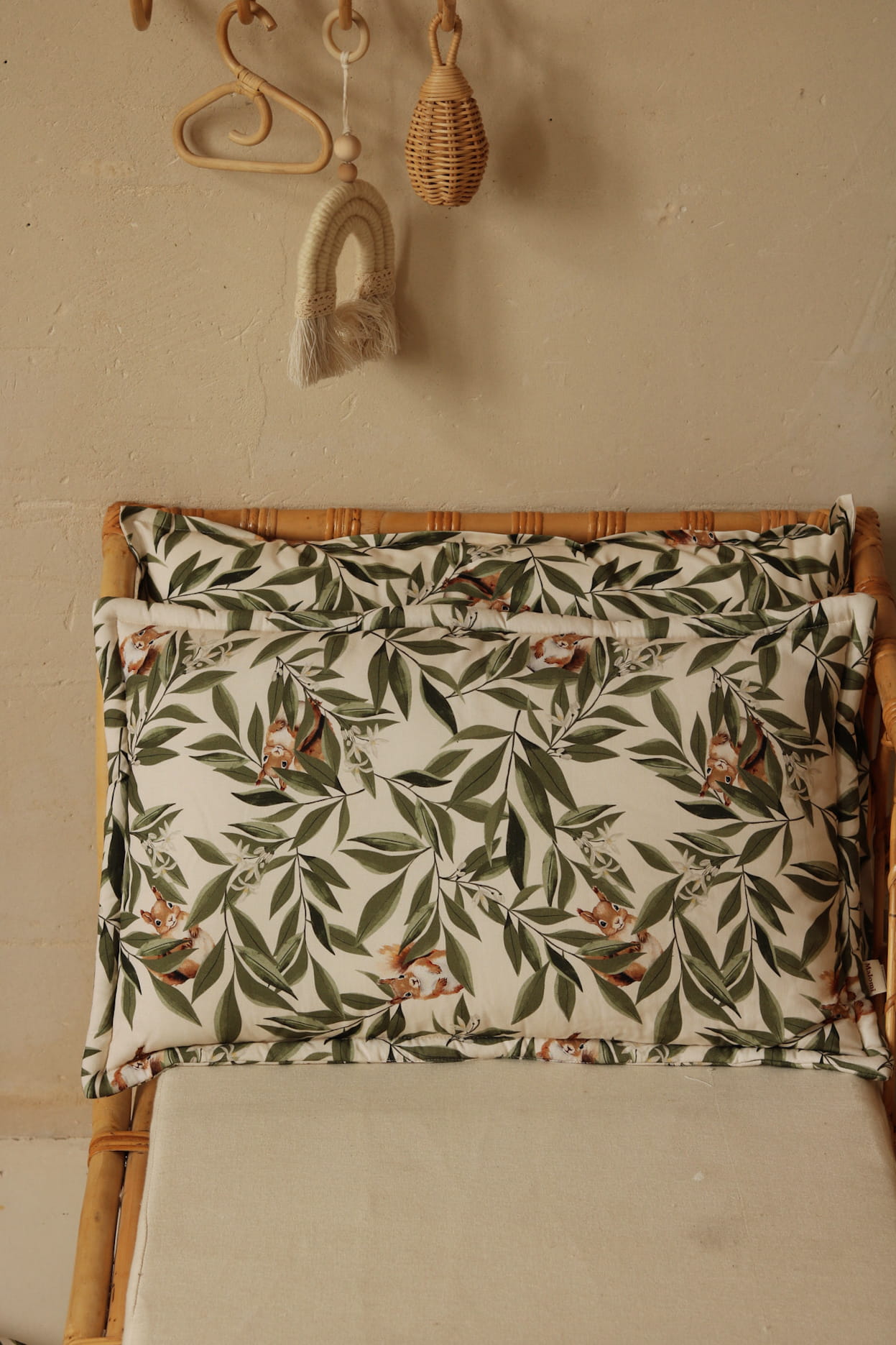 Kussen en dekentje met bladeren- en eekhoornprint op een rotan kinderbankje, tegen een beige muur met natuurlijke decoraties zoals een regenbooghanger en rotan accessoires.