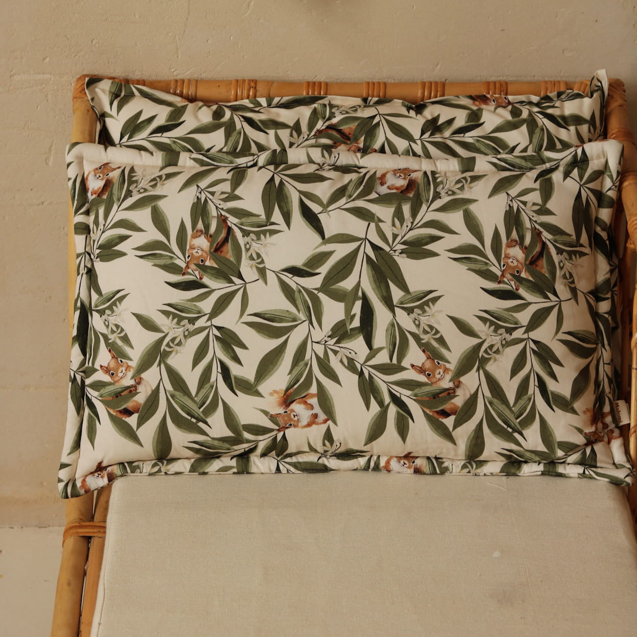 Kussen en dekentje met bladeren- en eekhoornprint op een rotan kinderbankje, tegen een beige muur met natuurlijke decoraties zoals een regenbooghanger en rotan accessoires.