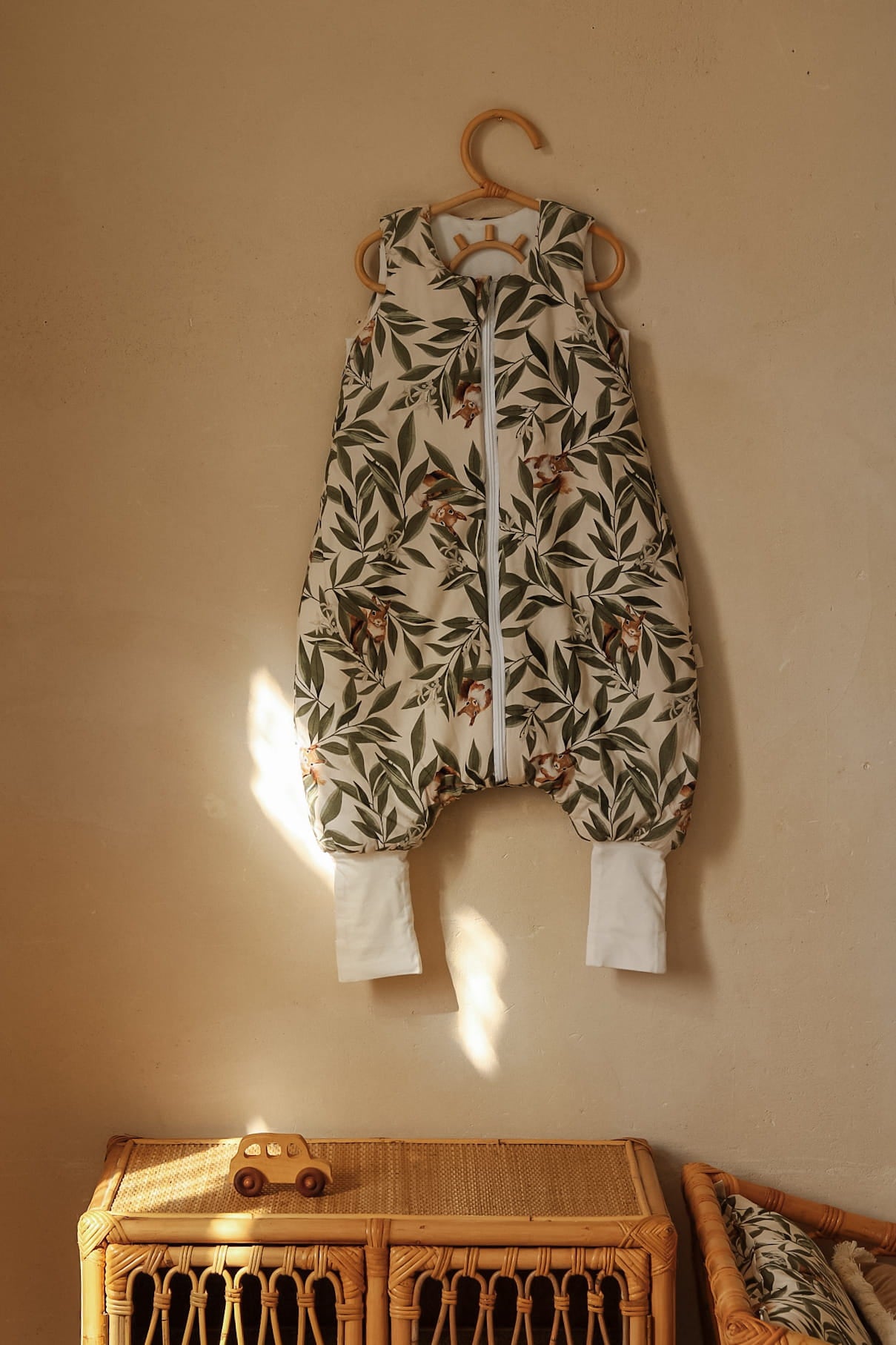 Slaapzak met woodland print hangt aan een rotanhaak tegen een beige muur. Op de voorgrond staat een rotankastje met een houten speelgoedautootje erop.